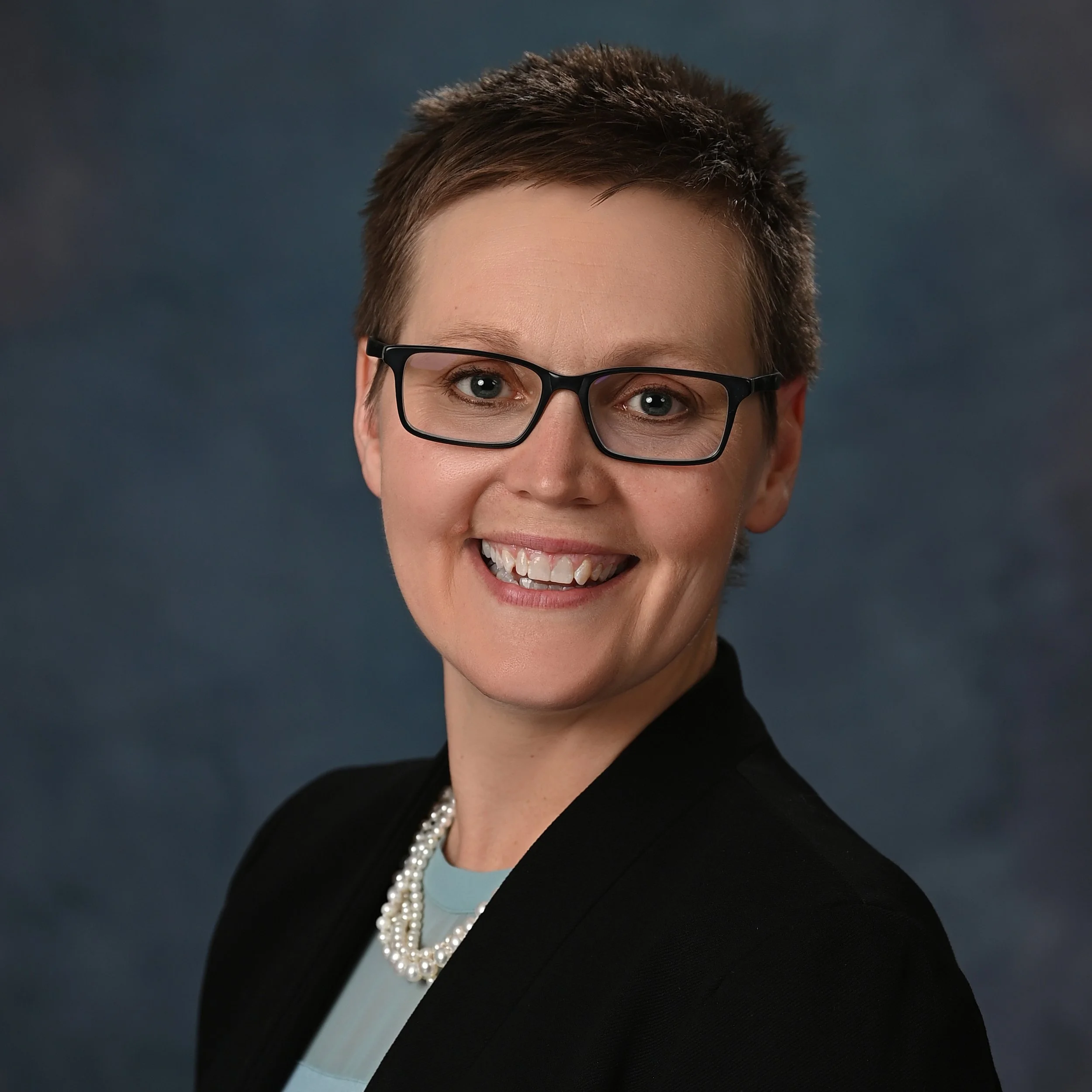 Dr. Erin Haugen wearing black glasses, a black jacket, a blue shirt, and pearls. She is smiling.
