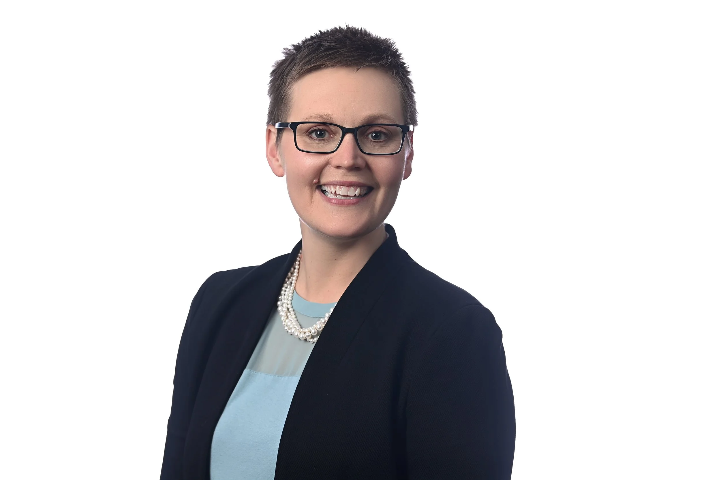 A woman with short hair and glasses wearing a navy blazer, a light blue top, and a pearl necklace, smiling at the camera against a white background.
