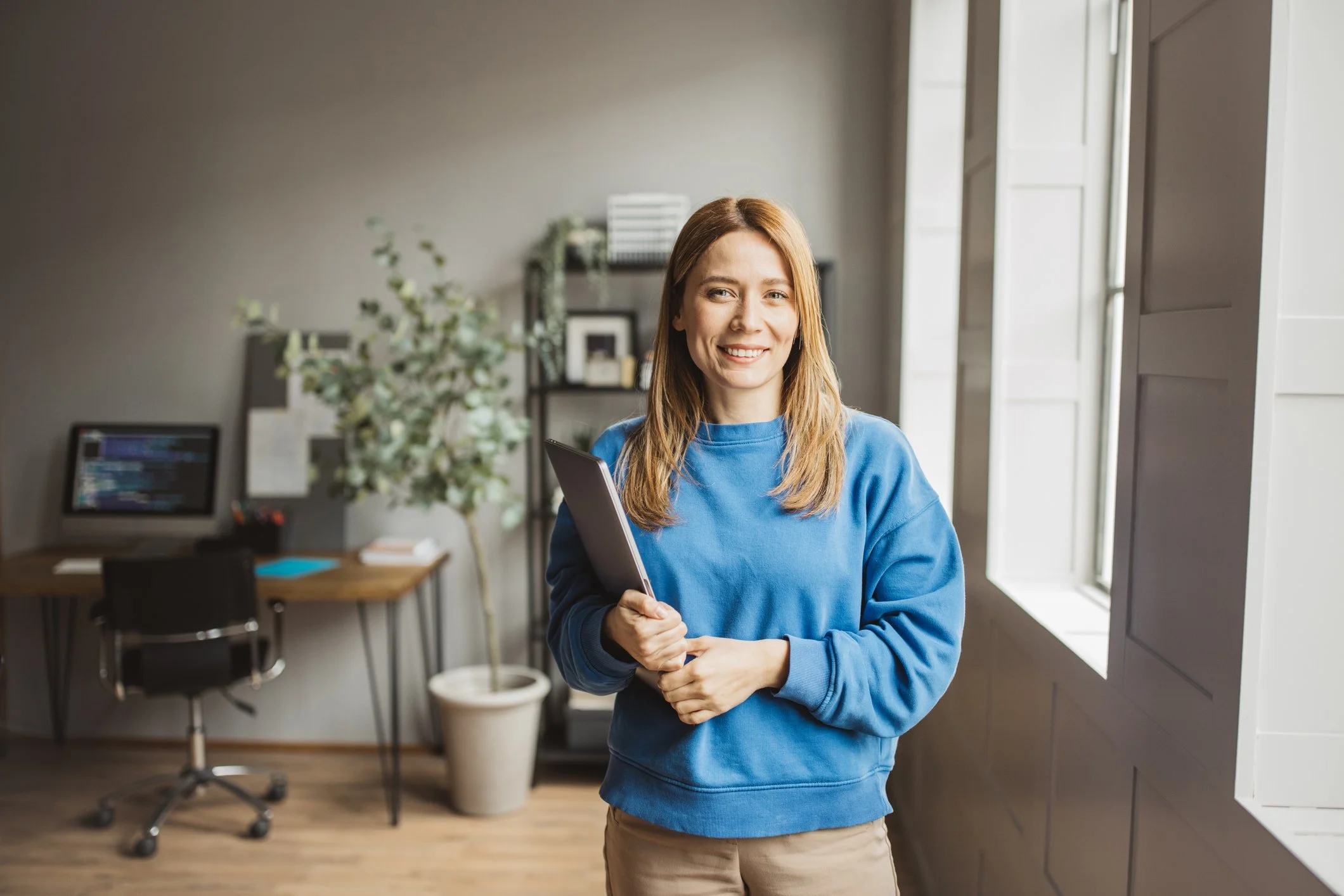 Young woman with long hair smiling and holding a tablet in a modern office space with a desk, chair, computer monitor, and large window.
