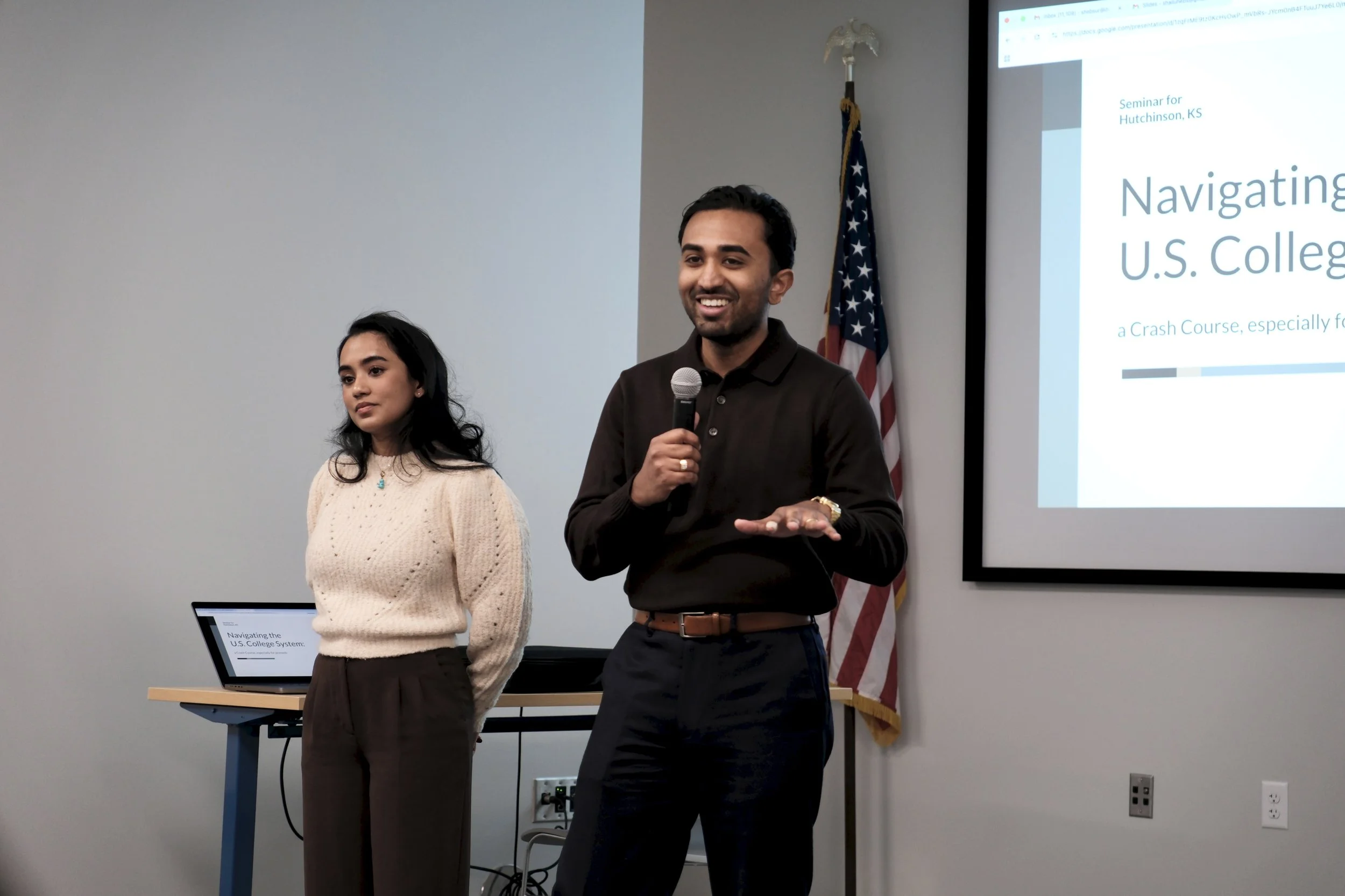 A man holding a microphone and smiling during a presentation in a conference room with a woman standing next to him. The presentation slide on the screen reads "Navigating U.S. College System" and an American flag is in the background.