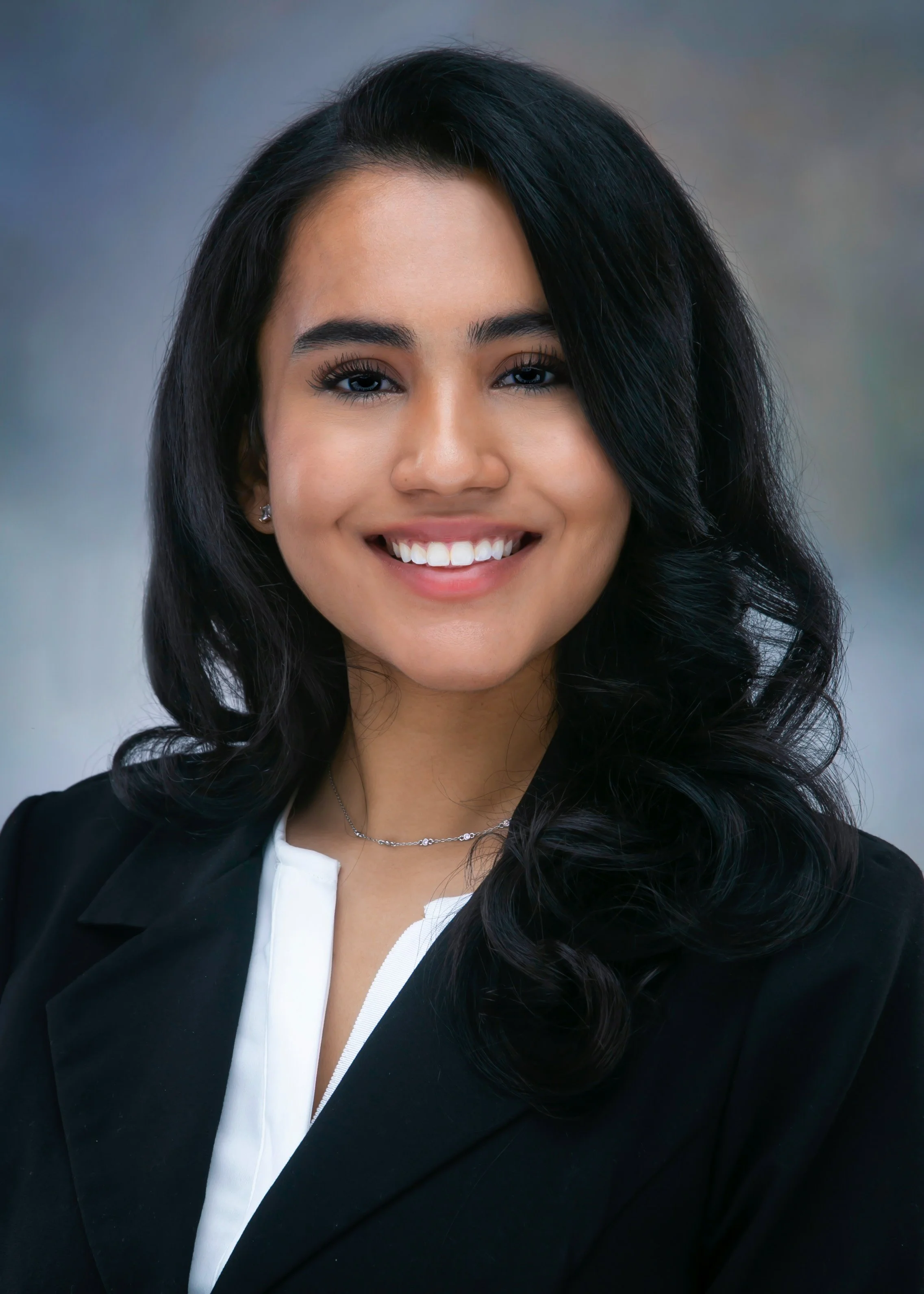 Professional portrait of a young female physician with long black hair, smiling, wearing a dark blazer and a white shirt, with a blurred background.