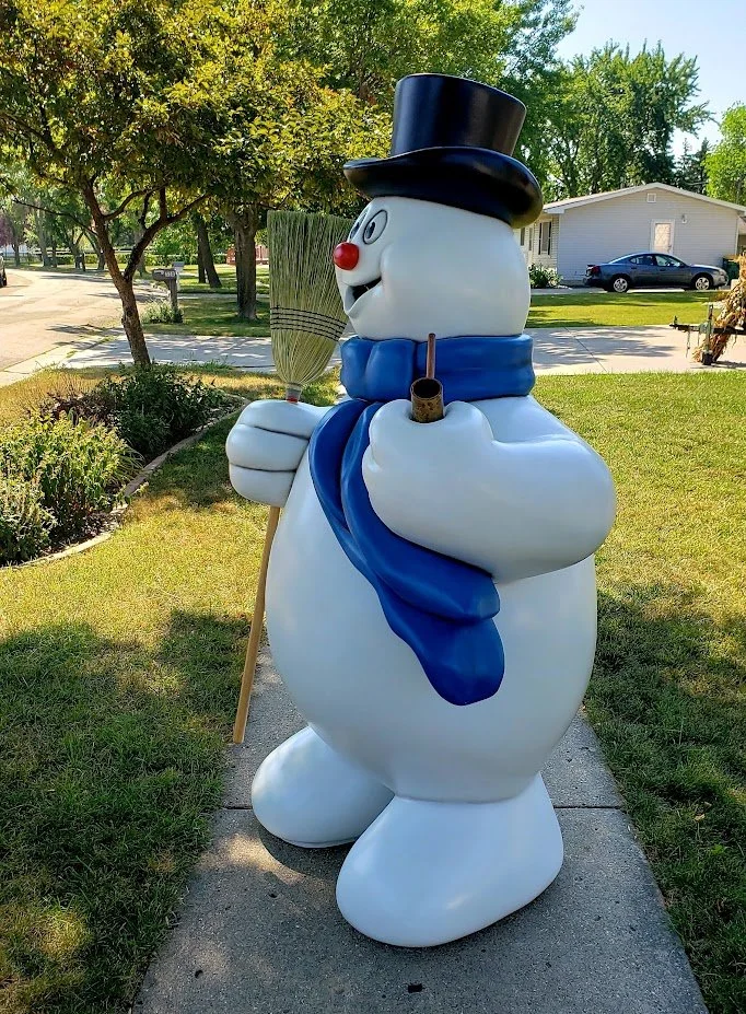 A snowman statue wearing a black top hat, holding a broom and a pipe, with a blue scarf around its neck, placed on a sidewalk in a suburban neighborhood.