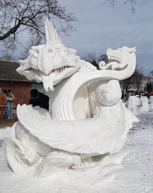 Snow sculpture of a dragon with a large head, open mouth, and detailed scales, built outdoors in a snowy area.