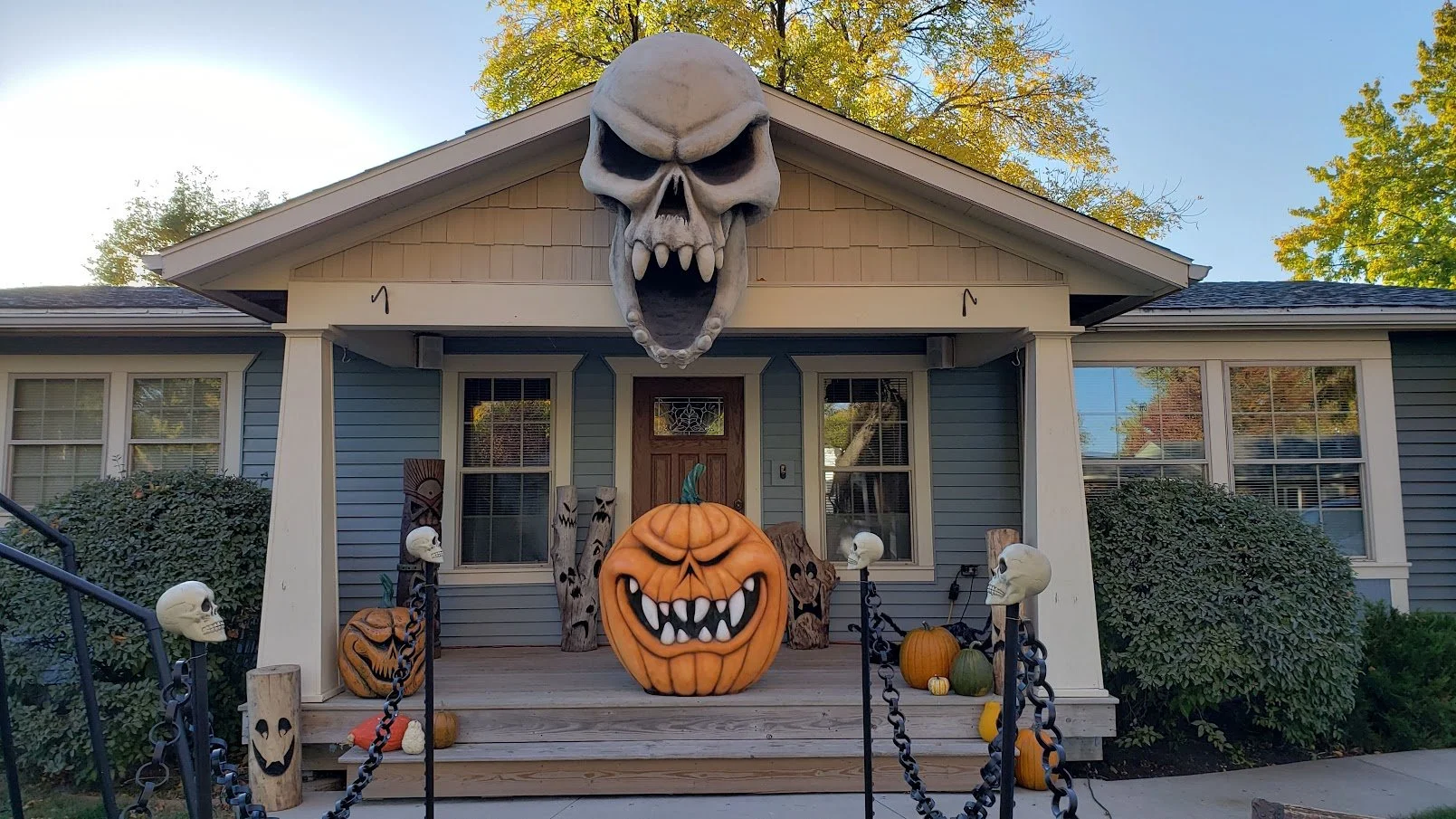 House decorated for Halloween with a large hanging skull above the porch, a carved pumpkin with a menacing face in the center, and various smaller pumpkins and skull decorations around the porch.