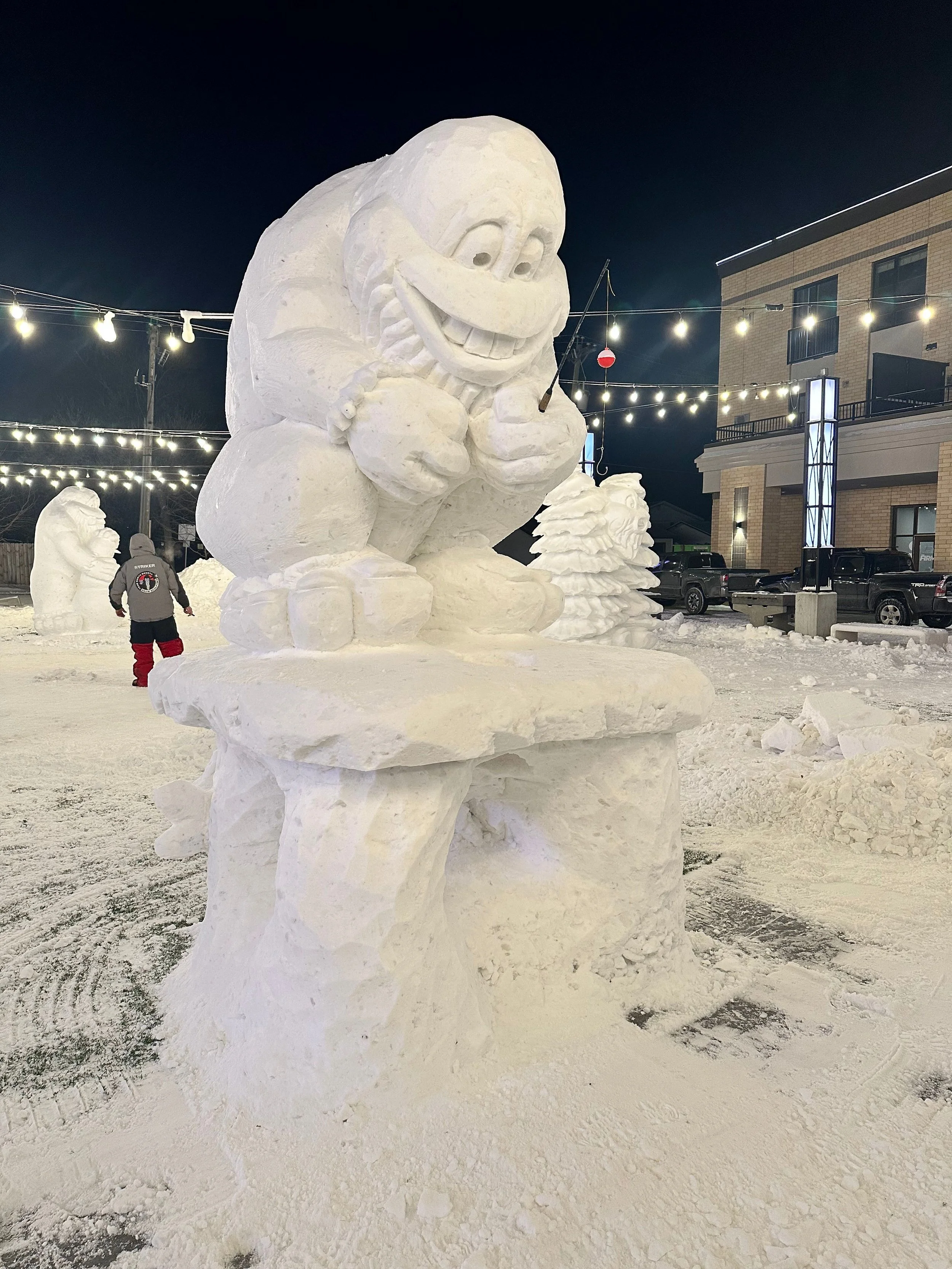 Snow sculpture of a cartoonish character with a smiling face, holding a fishing rod, and sitting at a table in an outdoor setting at night, with string lights overhead and a person walking in the background.