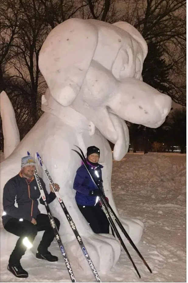 Two people posing with skis in front of a large snow sculpture of Snoopy sitting on his doghouse during winter night.