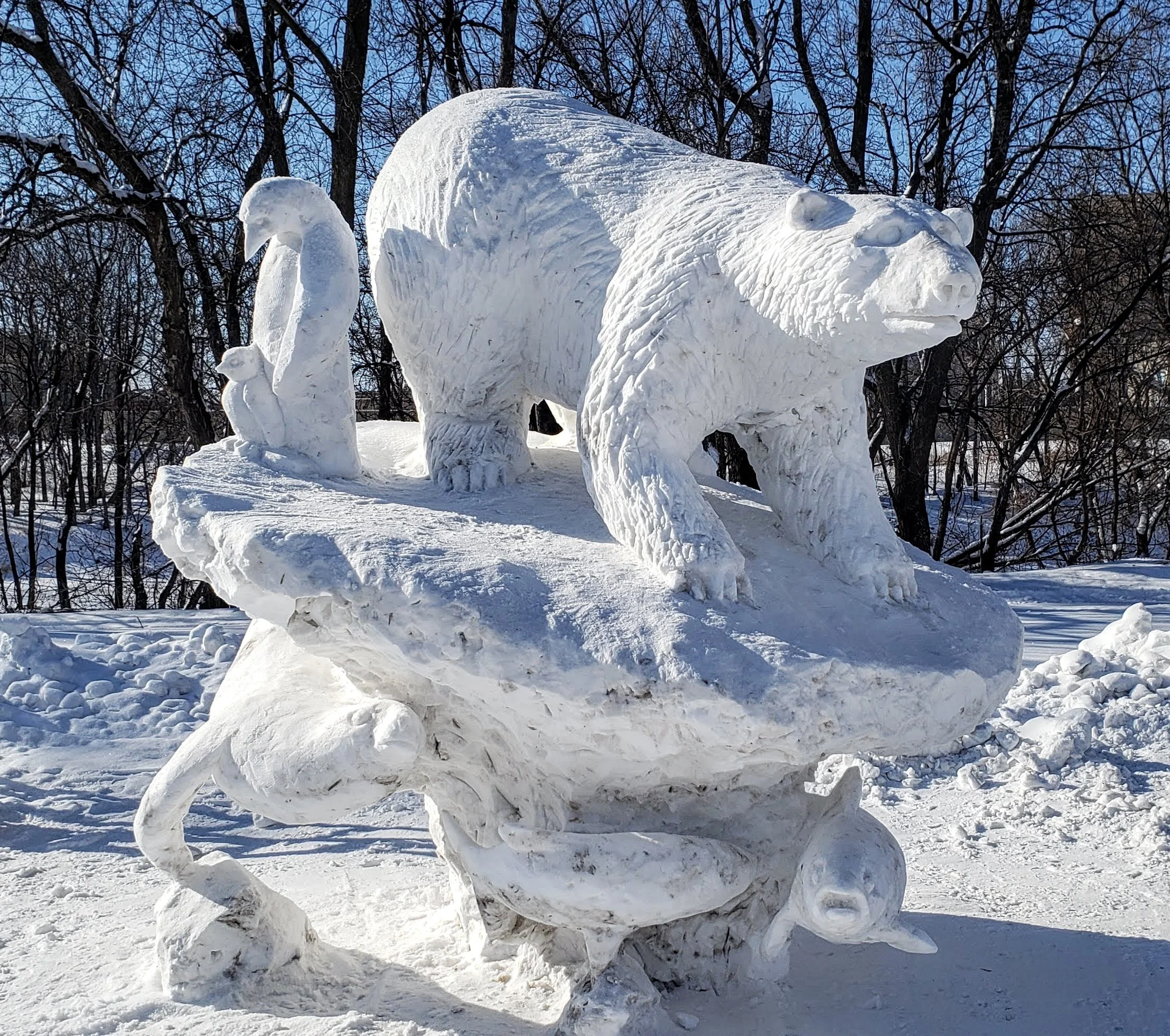 Snow sculpture of a bear walking on a rock, with a swan and a small bird on its back, set against a wintery backdrop of leafless trees.