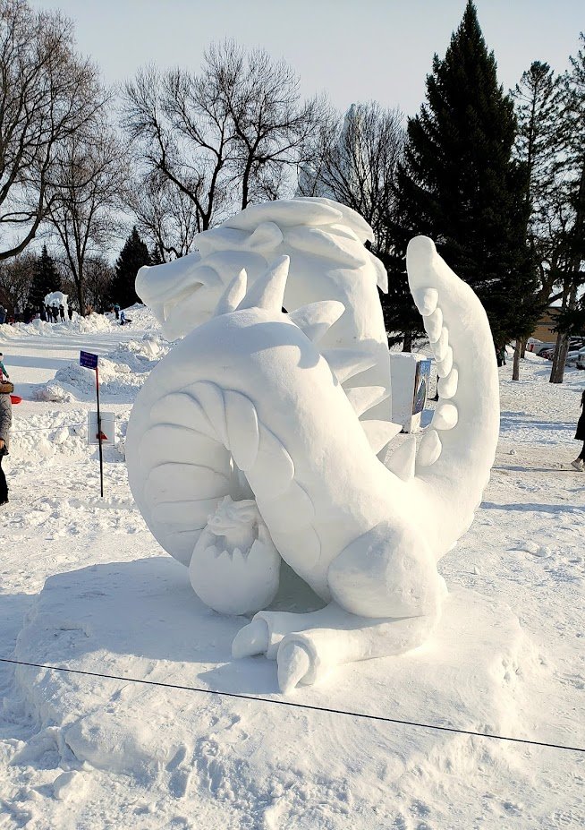 A large snow sculpture of a cartoon dragon with a fierce expression, sharp teeth, and a spiked tail, set outdoors in a snowy park with bare trees and a few people in the background.