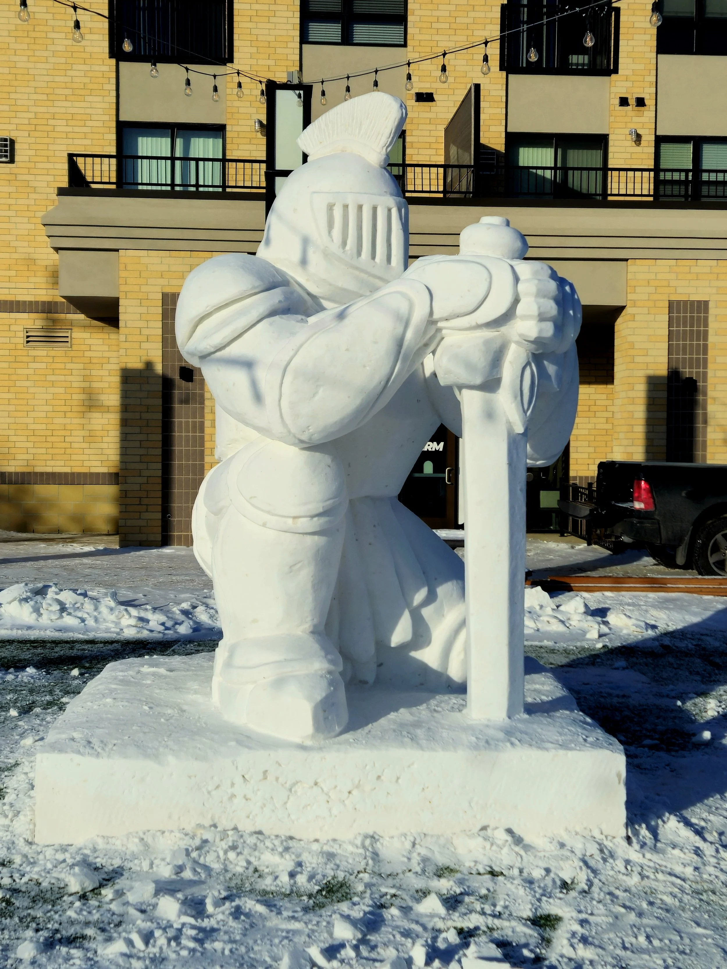 Snow sculpture of a medieval knight holding a large sword, with a helmet and armor, in front of a brick building and parked cars.