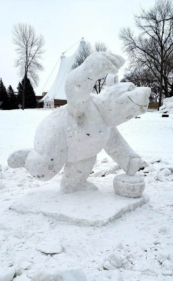 Snow sculpture of a cartoonish animal, possibly a dog or bear, balancing on a ball, outdoors in a snowy area with leafless trees in the background.