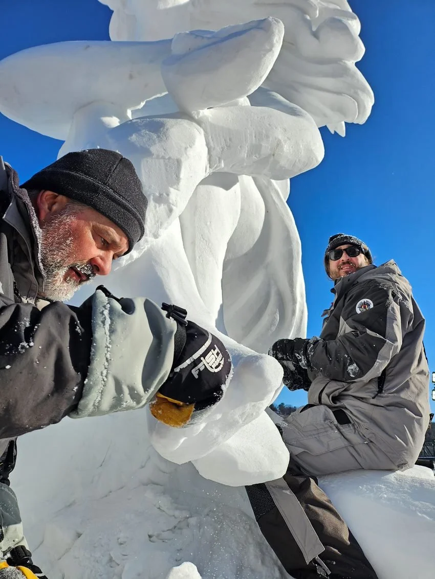 Two men in winter gear sculpting or carving a large snow sculpture outdoors on a bright, sunny day.