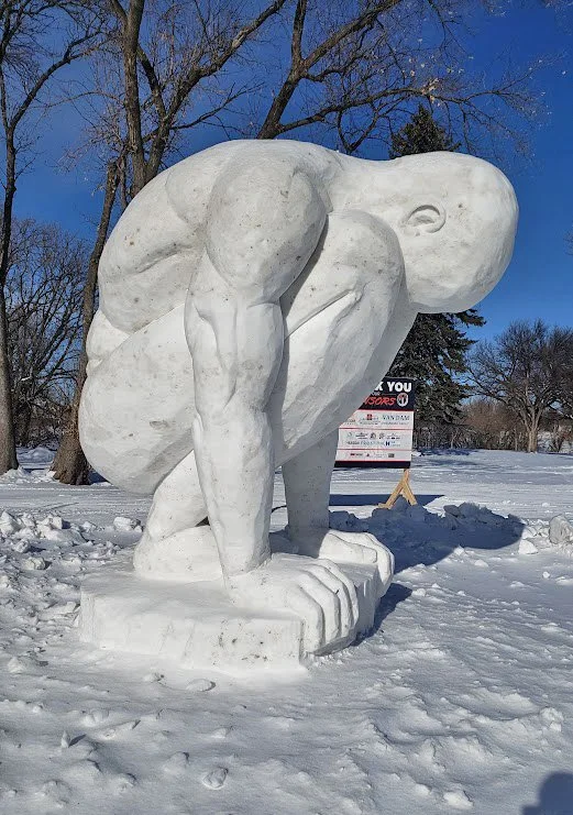Snow sculpture of a crouching person with bent knees and arms wrapped around their legs, outdoors on snow-covered ground with leafless trees and a sign in the background.