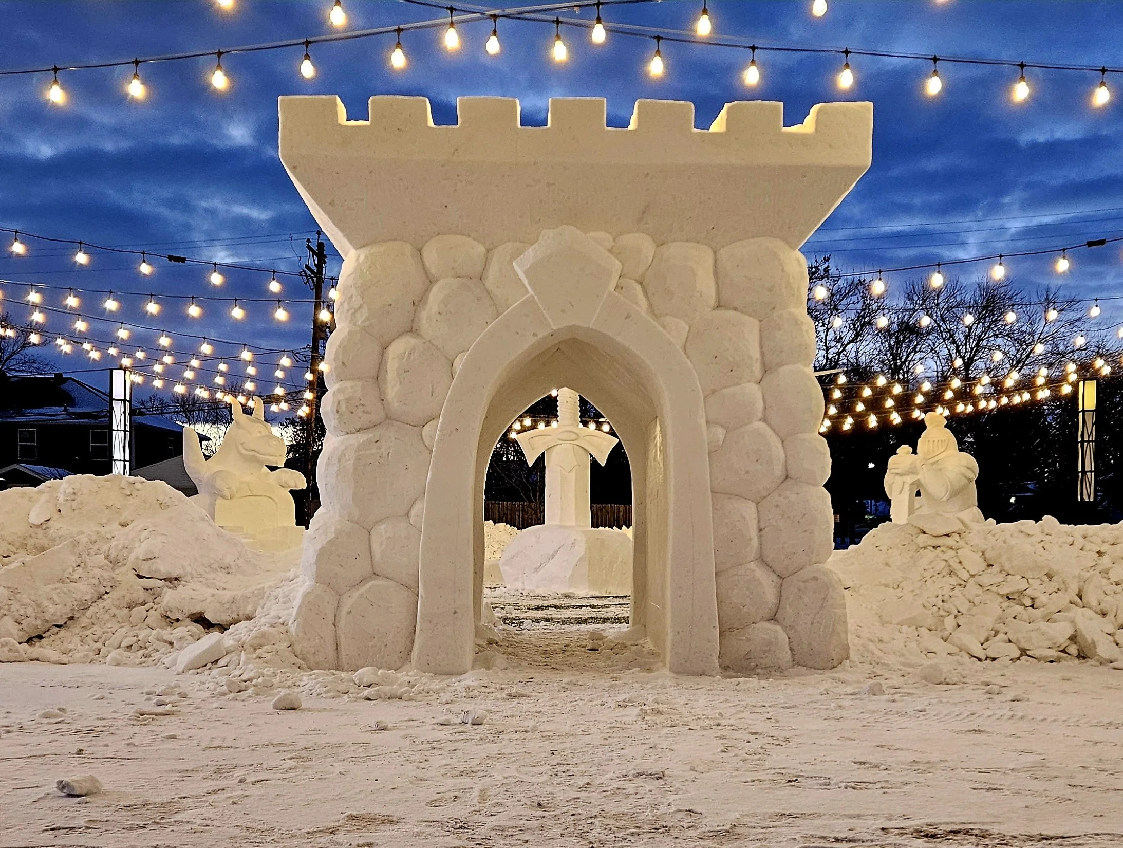 An outdoor snow sculpture of a castle gate with a sword in a shield behind it, flanked by snow sculptures of a unicorn and a knight. String lights hang above, and trees and houses are visible in the background at dusk.