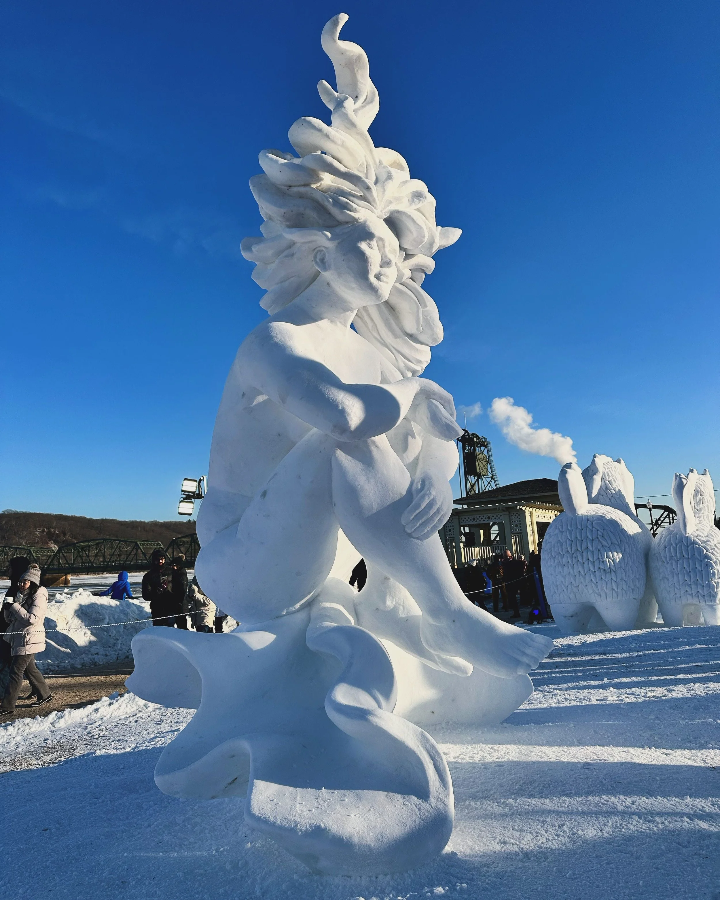 Large white snow sculpture of a mythological female figure with flowing hair and a serene expression, set outdoors in a snowy area on a clear day.