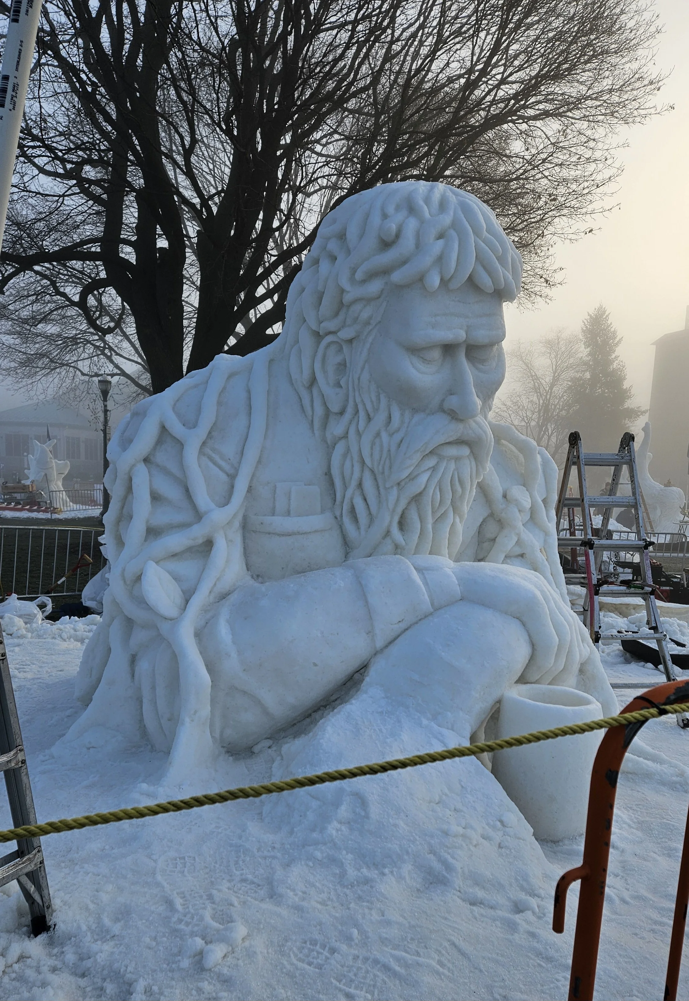A large snow sculpture of a bearded man with long hair, sitting with arms crossed, outdoors in a snowy park.