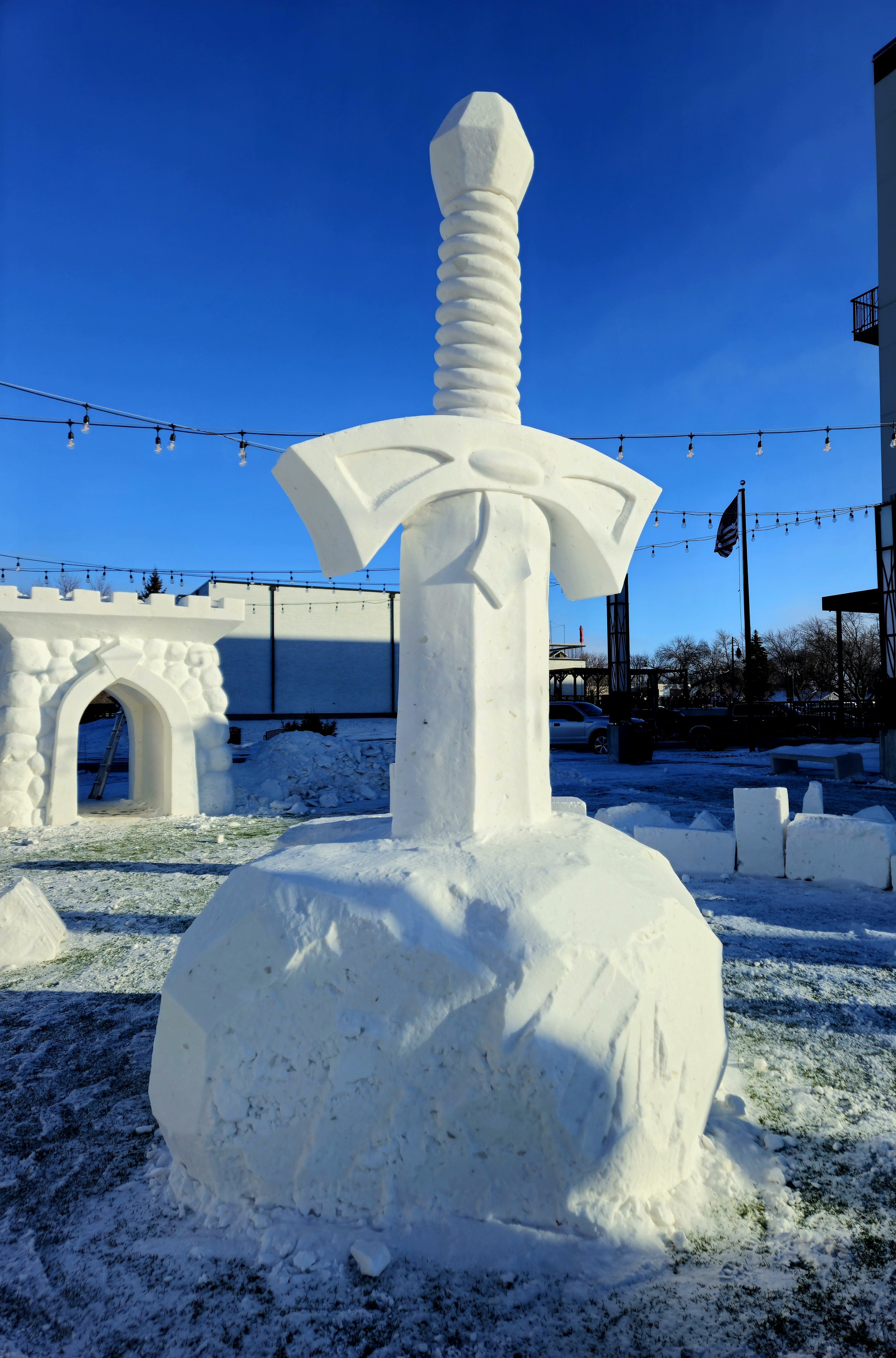 Snow sculpture of a large sword embedded in a snowball, set outdoors on a clear winter day with a blue sky.
