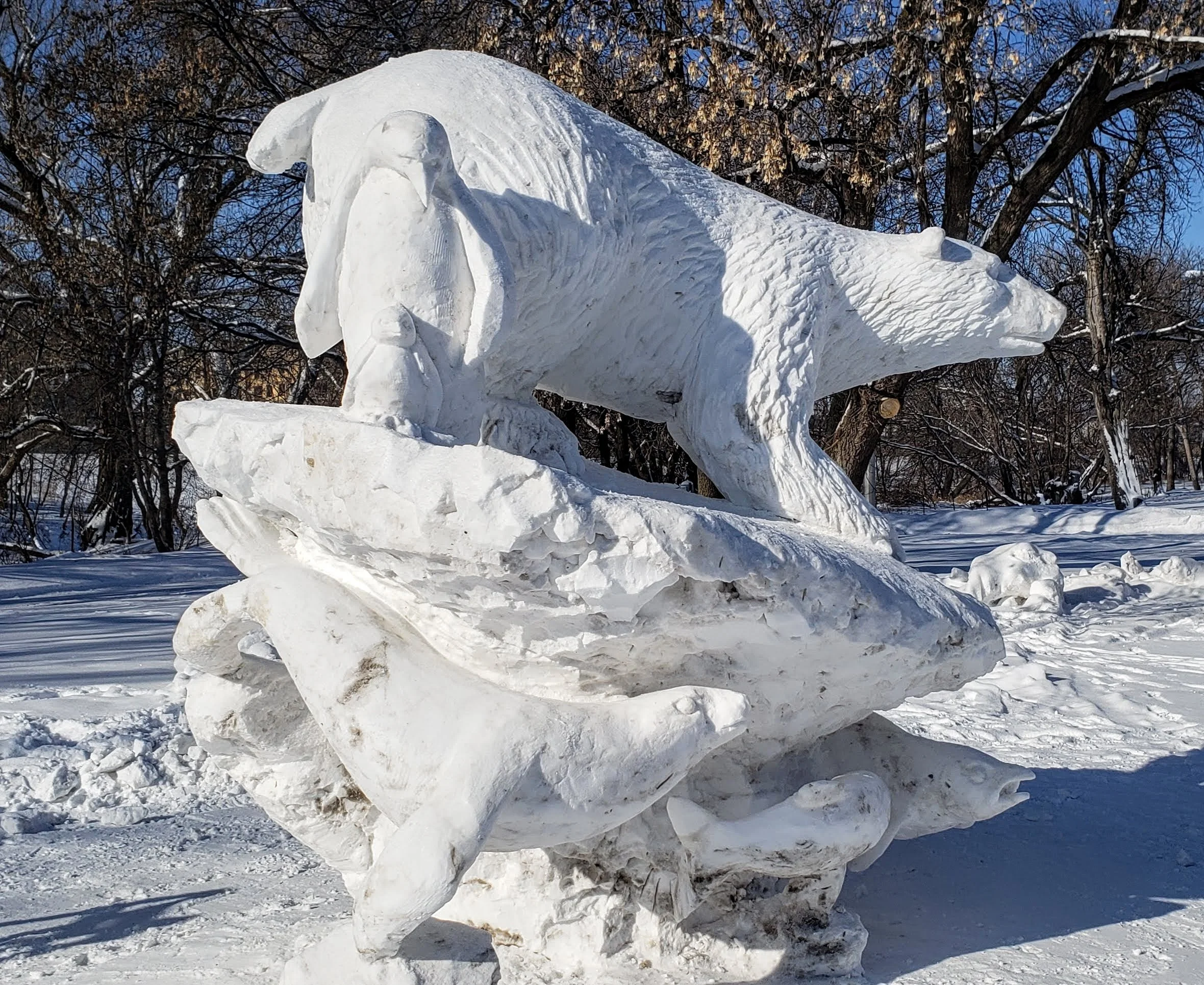Ice sculpture of a wolf in a snowy outdoor setting with leafless trees in the background.
