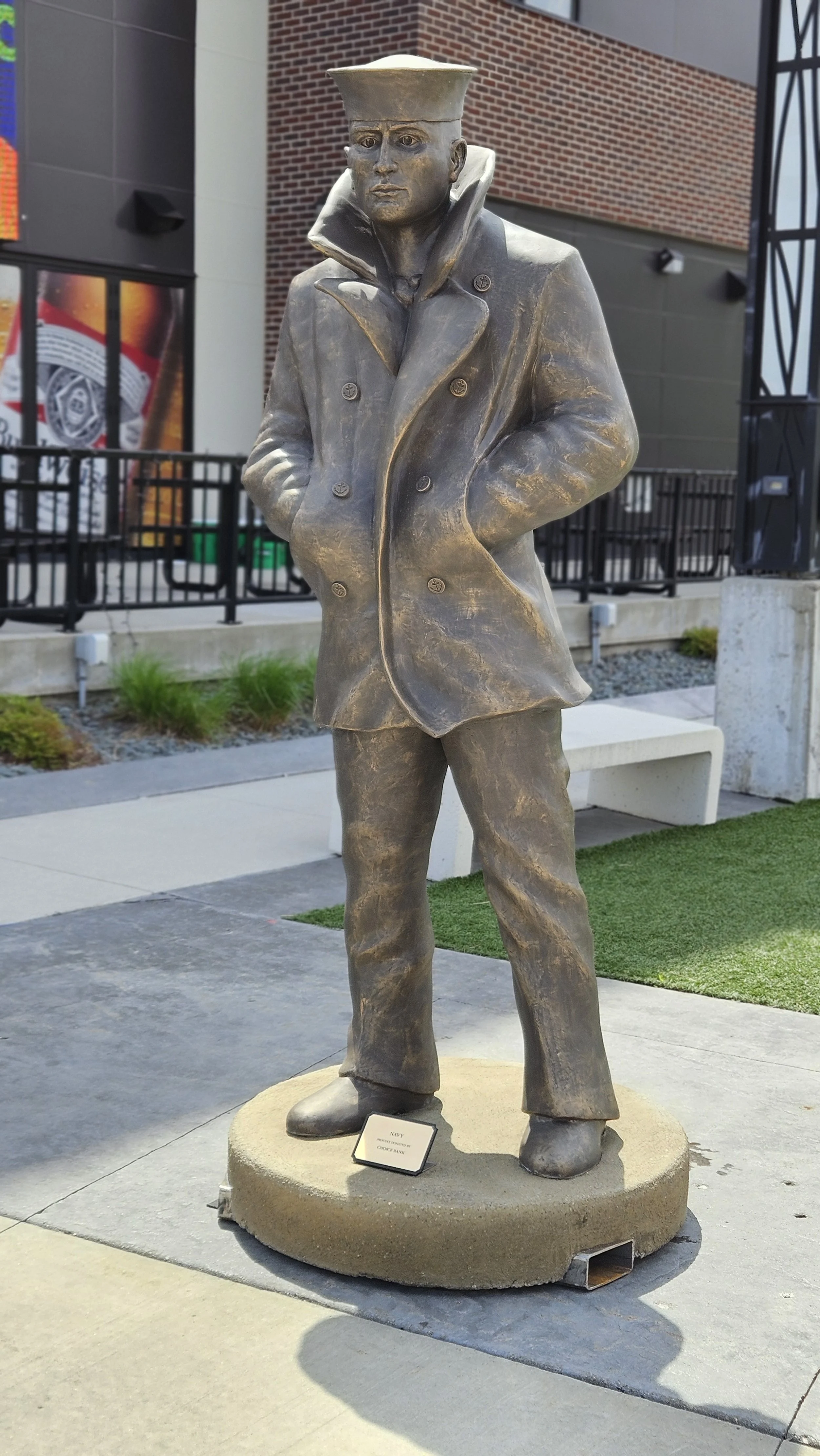 Bronze statue of a man wearing a coat and hat, standing with hands in pockets, outdoors near a building.