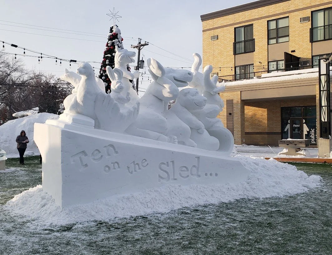 Snow sculpture of various animals on a sled with the words "Ten on the sled..." carved in the snow, with a decorated Christmas tree and buildings in the background.
