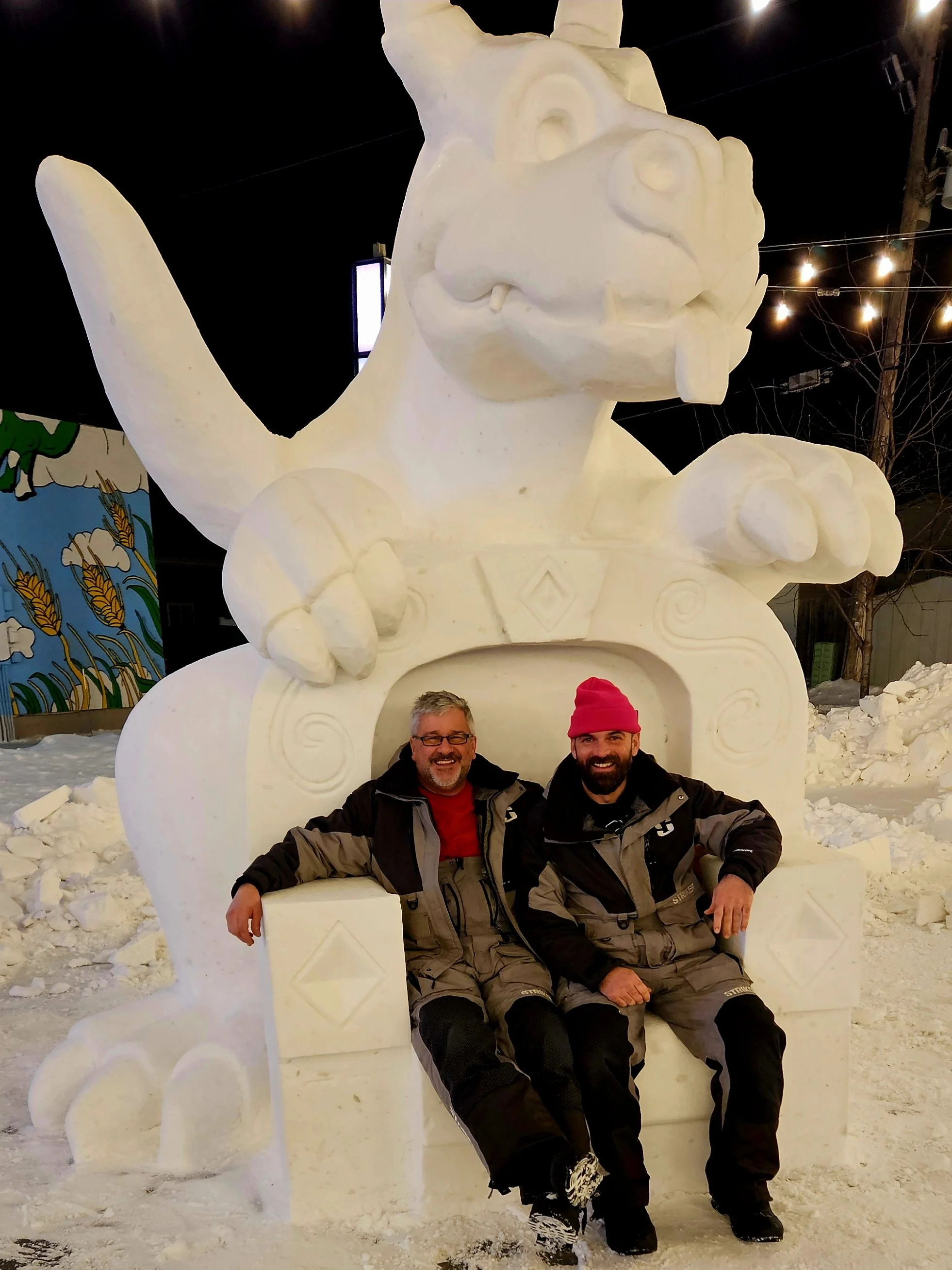 Two men sitting on a snow sculpture of a creature with a large head, big eyes, and a wide smile, in a snowy outdoor setting at night.