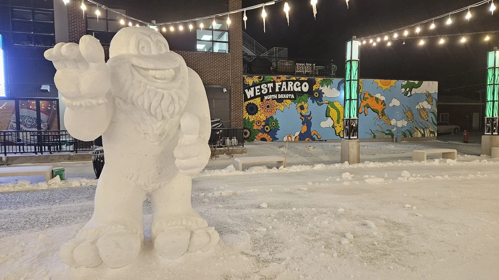 A large snow sculpture of a cheerful bearded man with a hat, waving, situated outdoors at night with festive string lights overhead. In the background, there is a mural on a wall that reads 'West Fargo North Dakota,' decorated with colorful flowers, 