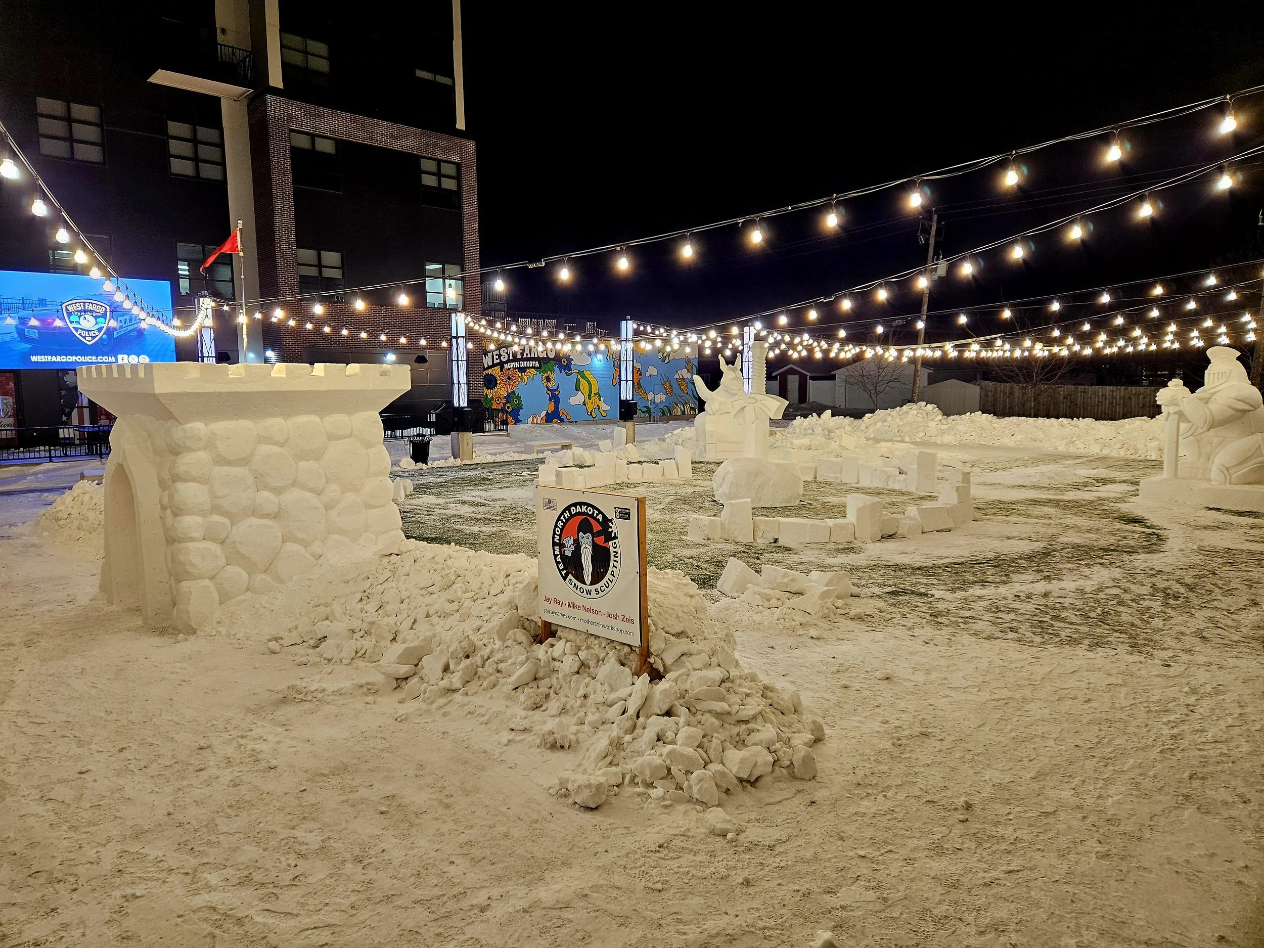 Nighttime view of an outdoor snow sculpture display with castles, characters, and other figures made of snow, illuminated by string lights, with a billboard and city buildings in the background.