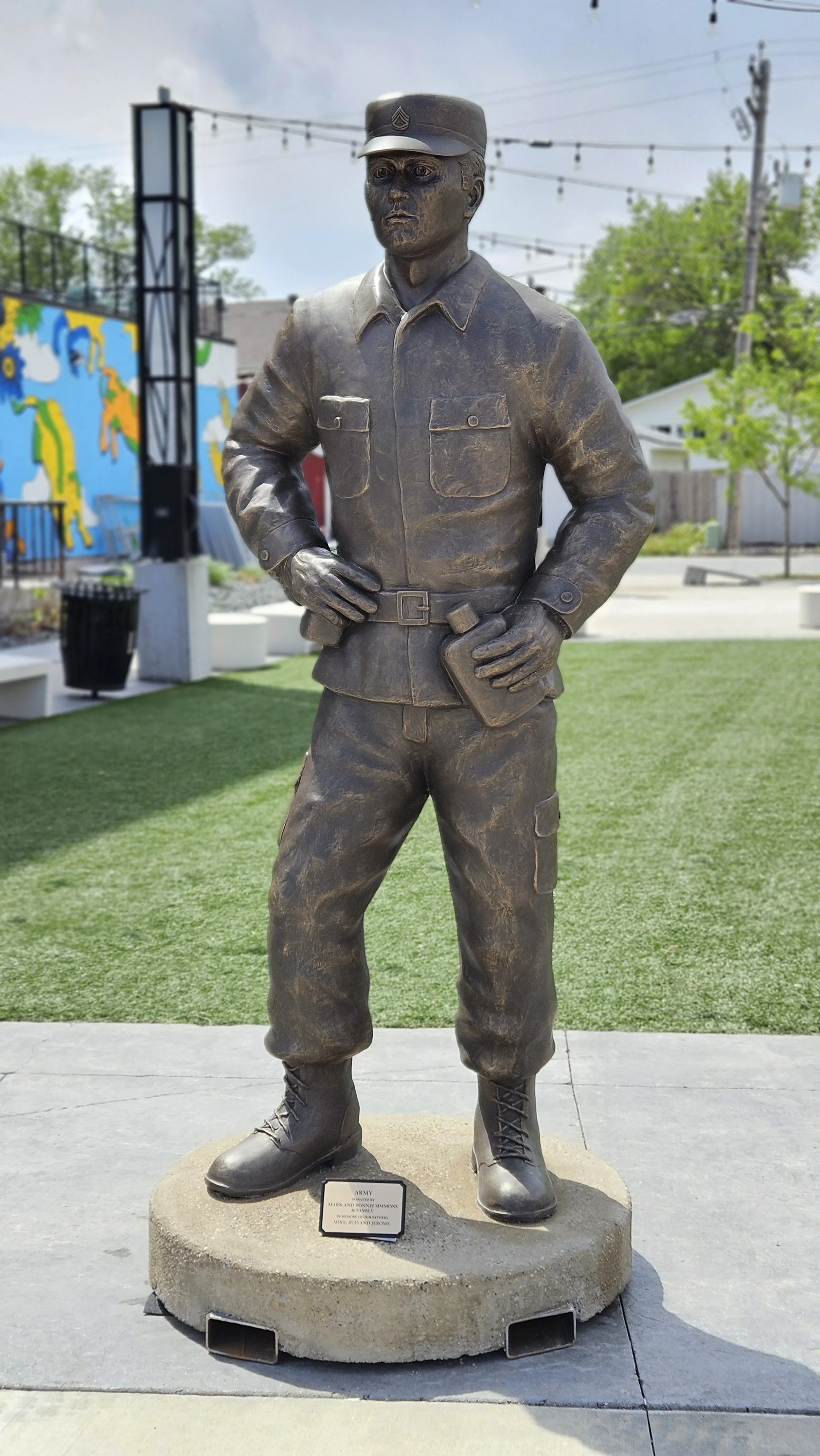Bronze statue of a soldier wearing a uniform with a cap, standing with hands on hips, holding a book in one hand, on a circular concrete base, in an outdoor park with greenery and a colorful mural in the background.