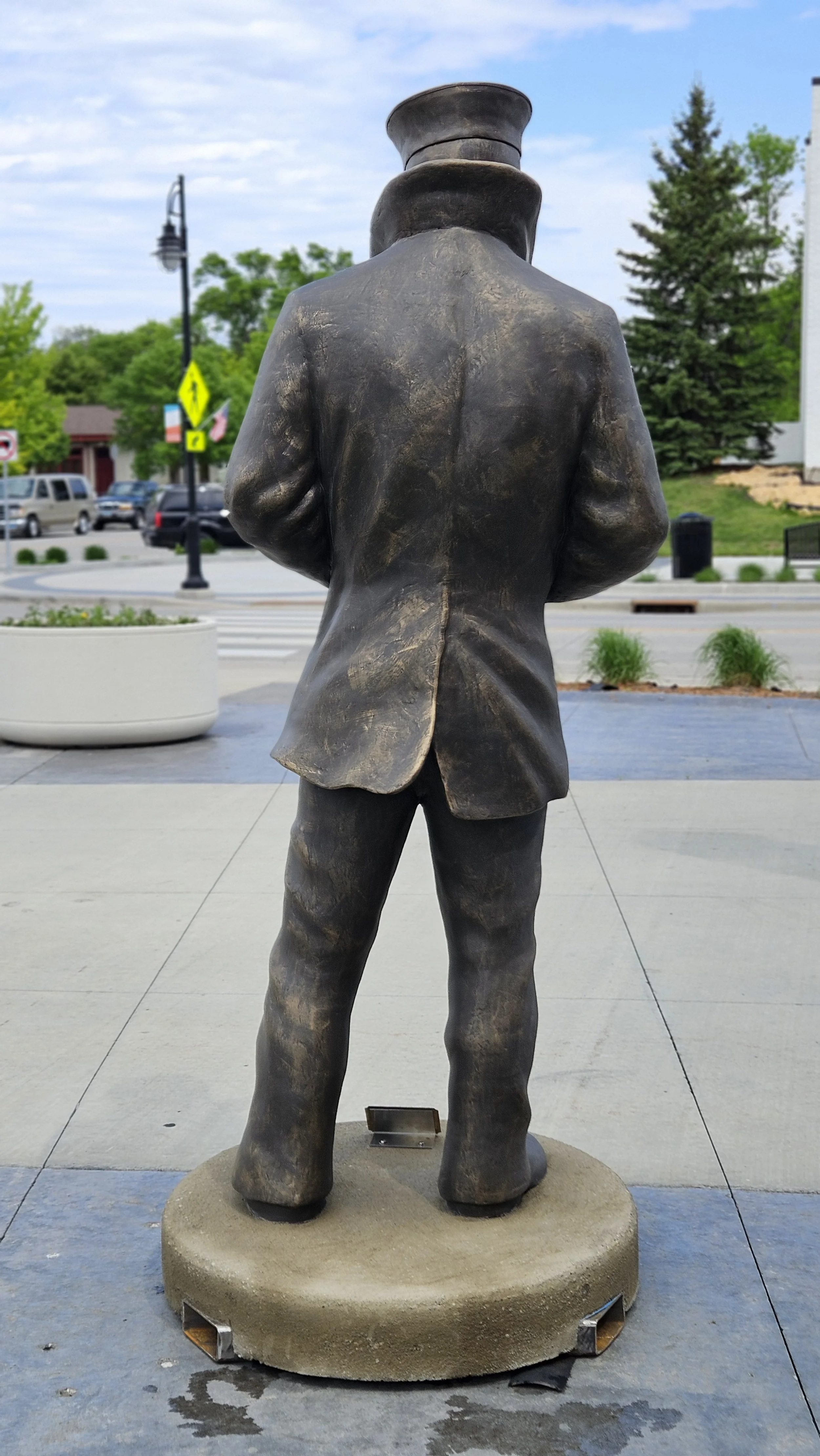 Bronze statue of a man in a suit and hat, standing with his back facing the camera, located outdoors on a circular base with a blurred background of trees, cars, and a street.