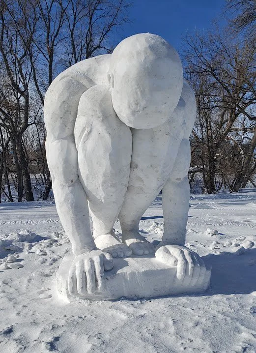 Large white snow sculpture of a person in a crouched position with their hands on the ground, set outdoors in a snowy landscape with leafless trees and a clear blue sky.