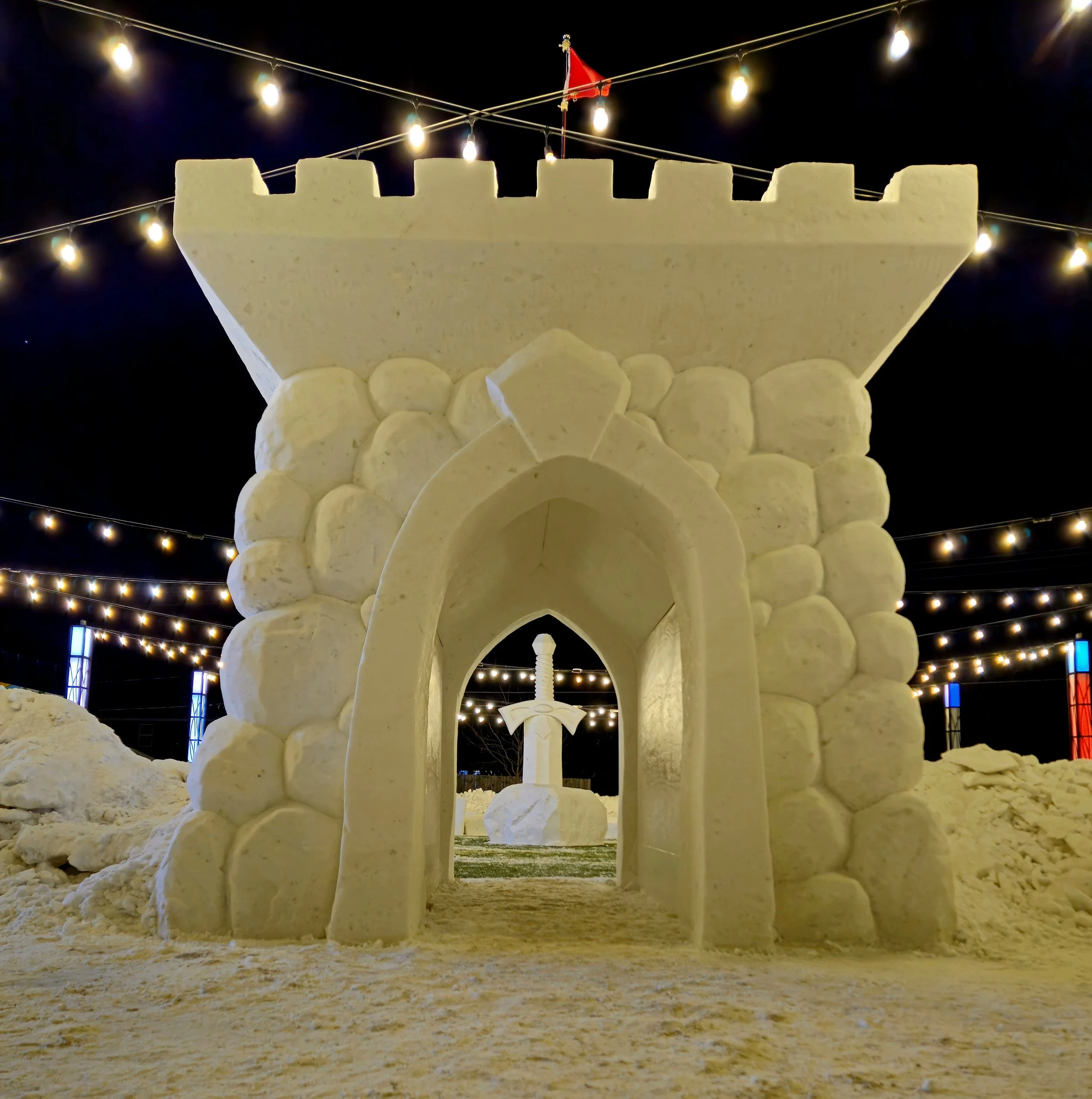 Sandcastle designed to resemble a castle with an archway and a sword sculpture inside, illuminated by string lights at night.