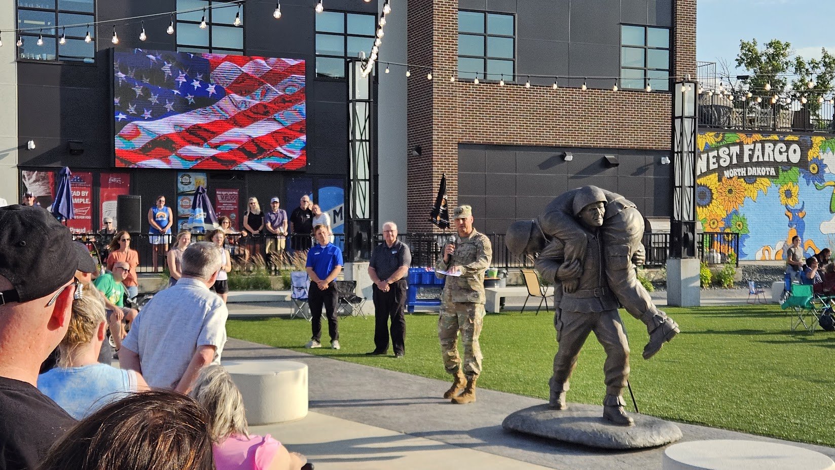A crowd gathers outdoors in front of a modern building with large screens, some people are speaking or listening, and a soldier in camouflage uniform stands holding a microphone. There is a life-size statue of a soldier carrying a wounded comrade on 