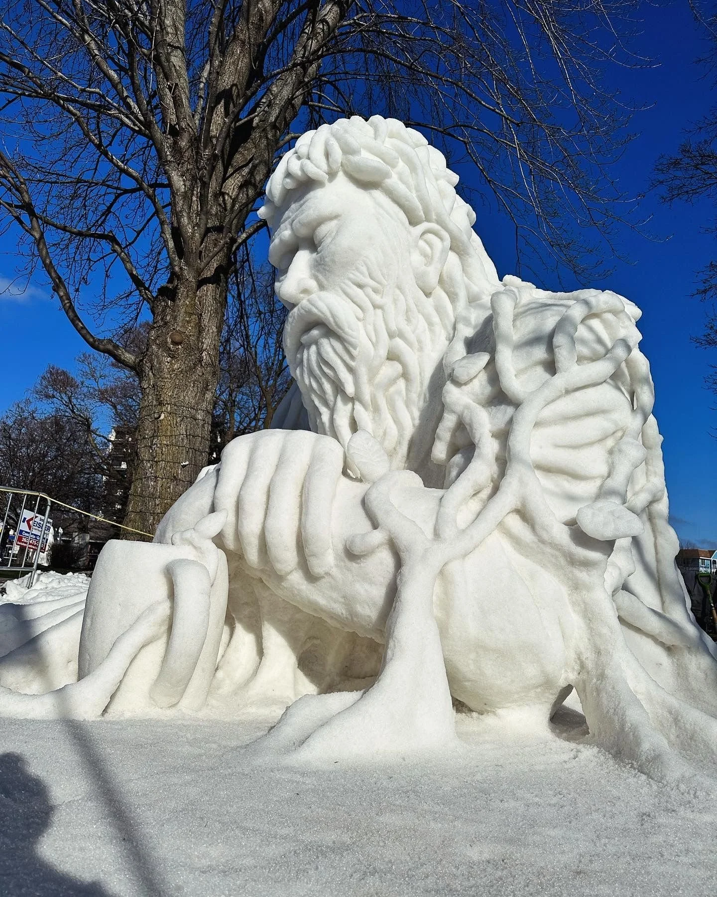 An intricate snow sculpture of a bearded man with a serious expression, surrounded by snow and leafless trees, under a bright blue sky.