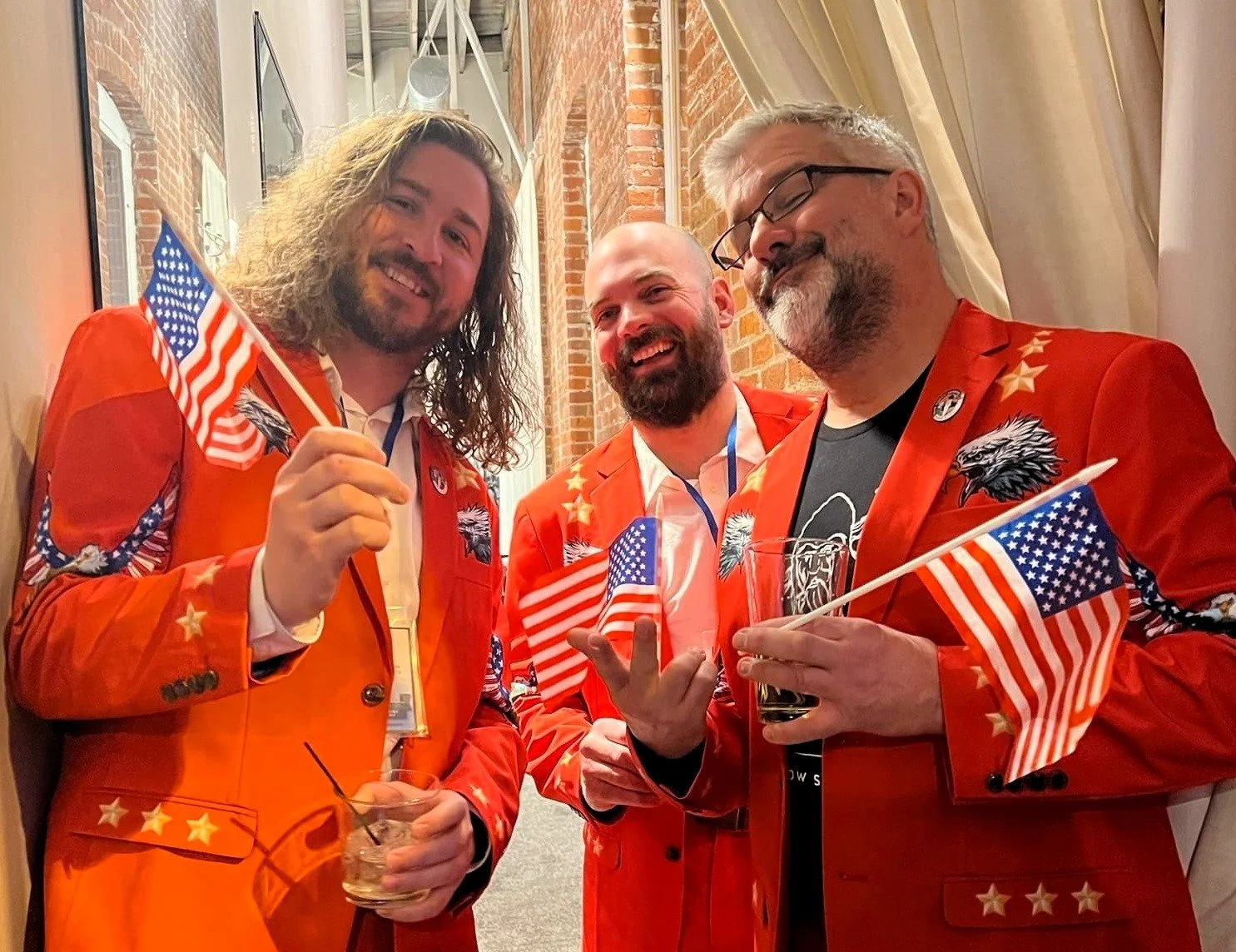 Three men dressed in red jackets with American flags, holding small American flags, smiling at a social gathering.