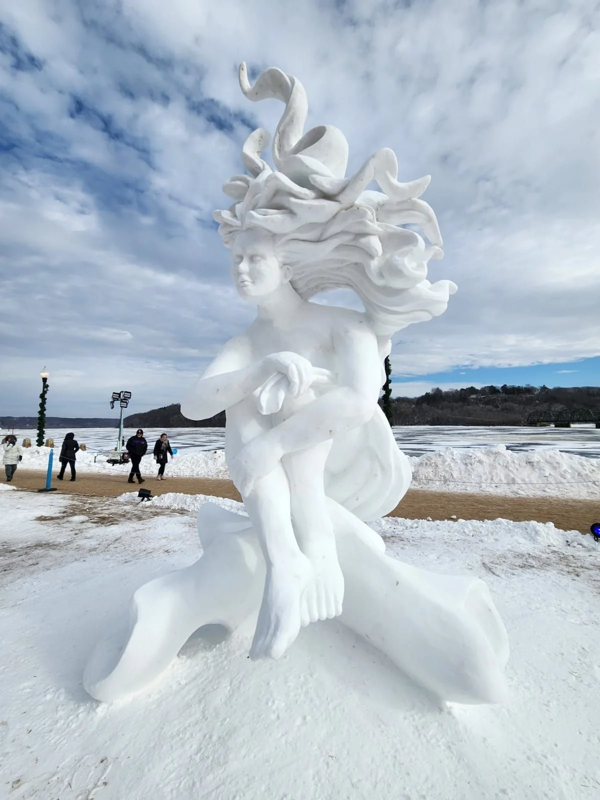 A large white sculpture of a woman with flowing hair intertwined with swans, kneeling on one knee in an outdoor snowy landscape with people walking in the background.