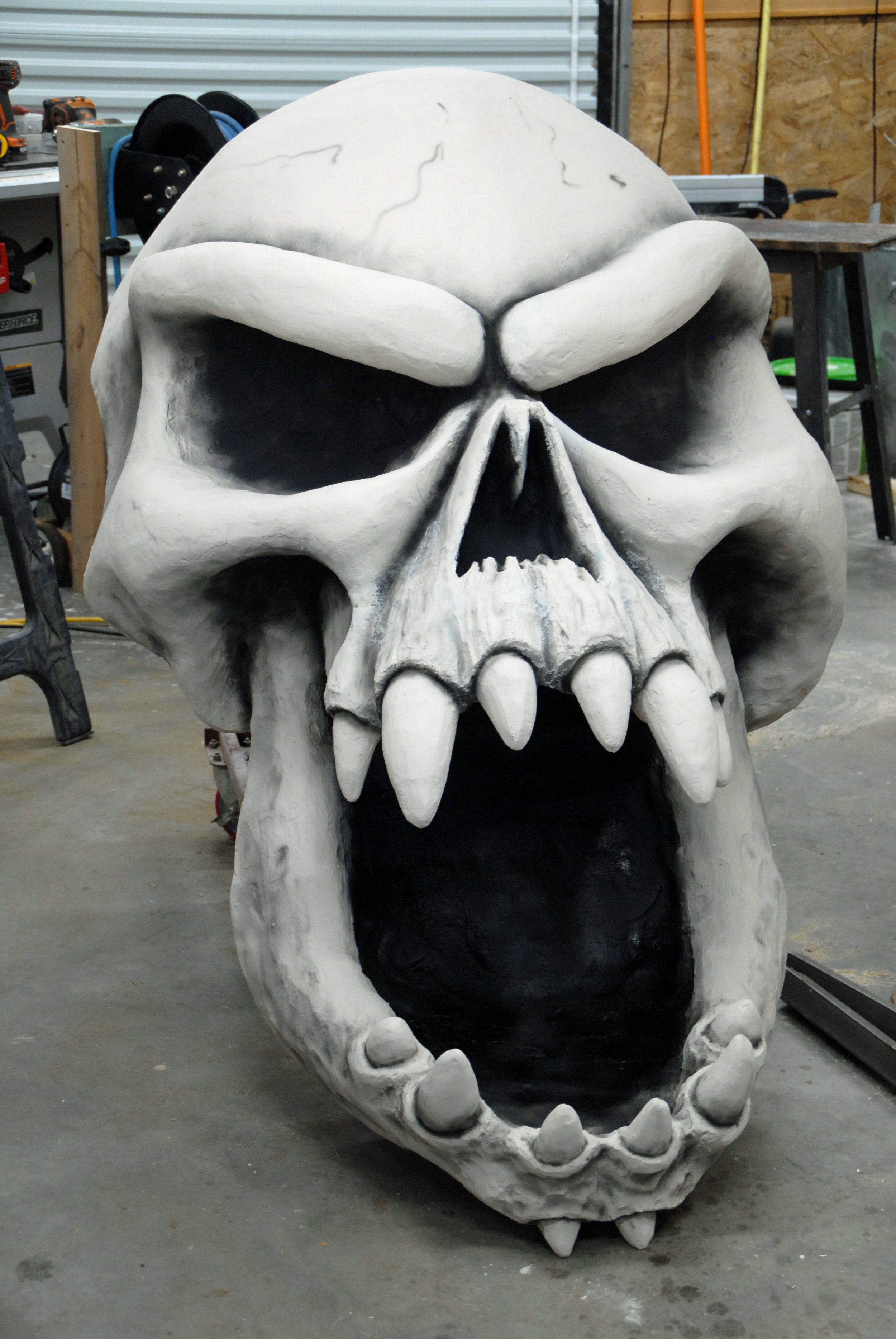 A large, detailed sculpture of a monstrous skull with exaggerated features including sharp teeth, prominent brow ridges, and hollow eye sockets, situated in a workshop.