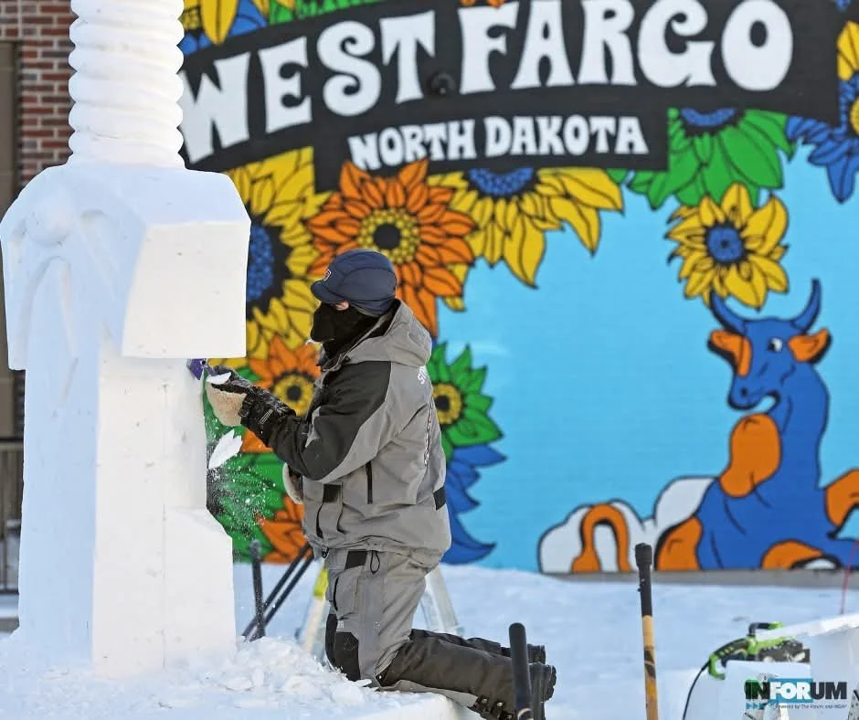 A person in winter gear carving a snow sculpture in front of a colorful mural that reads 'West Fargo North Dakota' with a blue background, sunflowers, and a blue and orange cow.