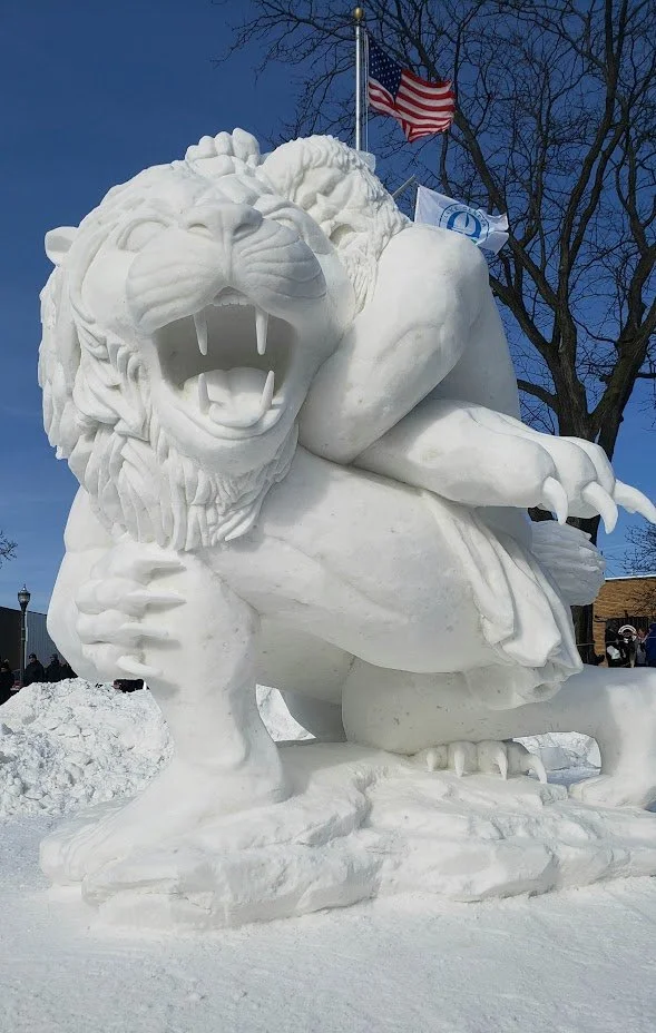A large snow sculpture of a roaring lion with a detailed mane, snarling face, and sharp claws, outdoors in winter under a clear blue sky with trees and American flags in the background.