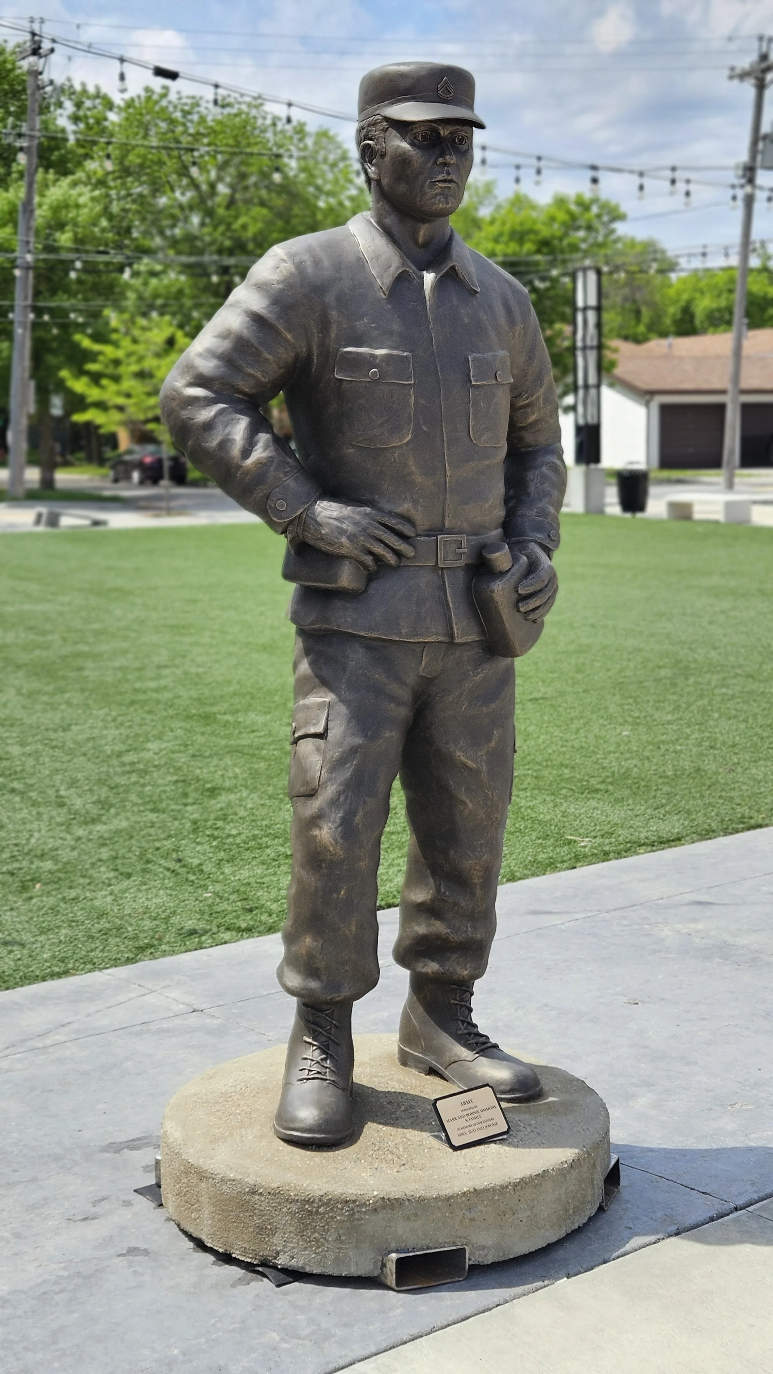 Bronze statue of a soldier in military uniform with hands on hips, standing on a circular cement base outdoors with grass and trees in the background.