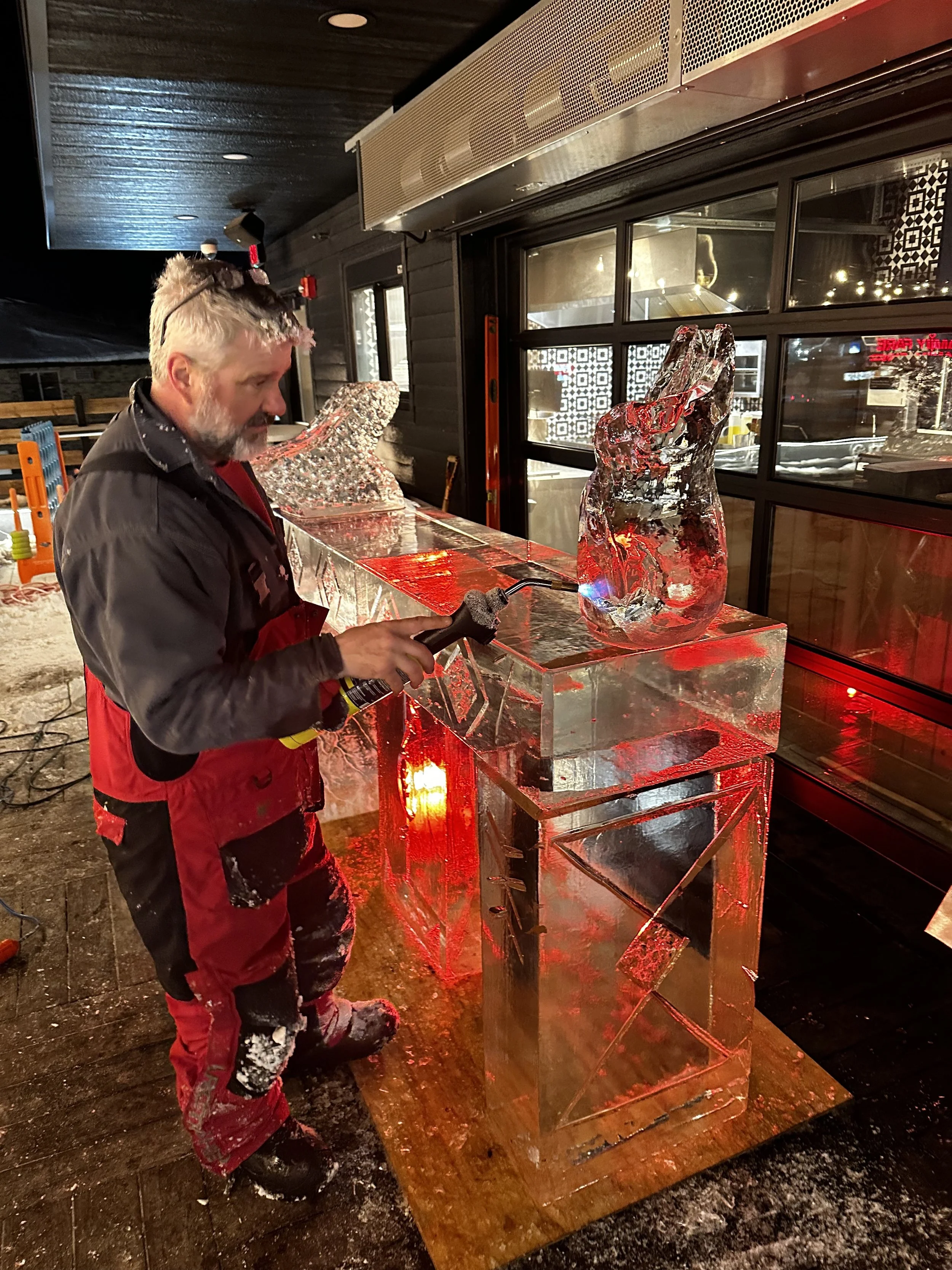 A man in a red and black snow suit is using a blowtorch to carve a large ice sculpture outside of a building at night. The sculpture resembles a vase or an abstract form and is illuminated with red lights.