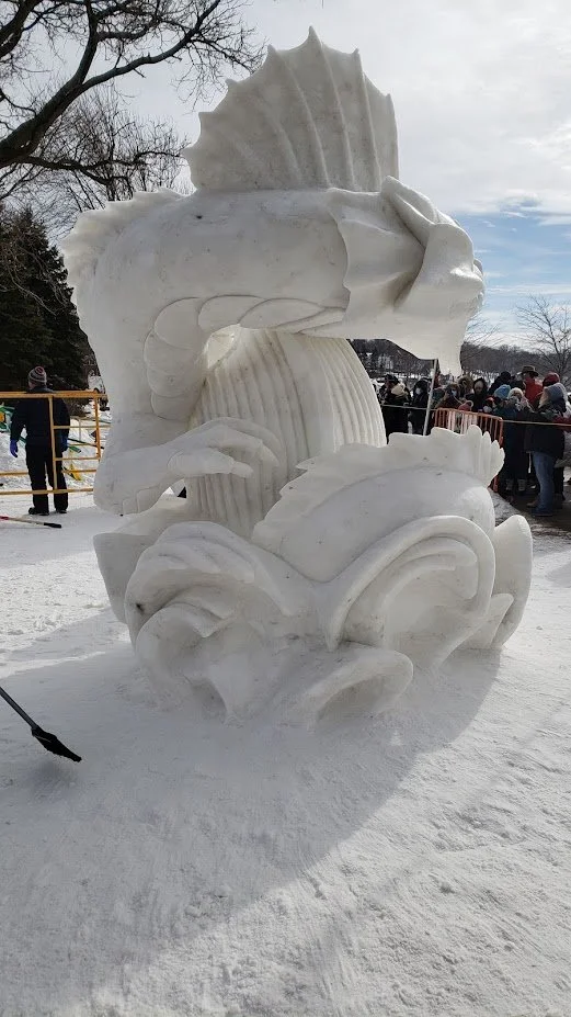 Large intricate snow sculpture of a dragon with detailed scales and features, set outdoors during winter with trees, people, and cloudy sky in the background.