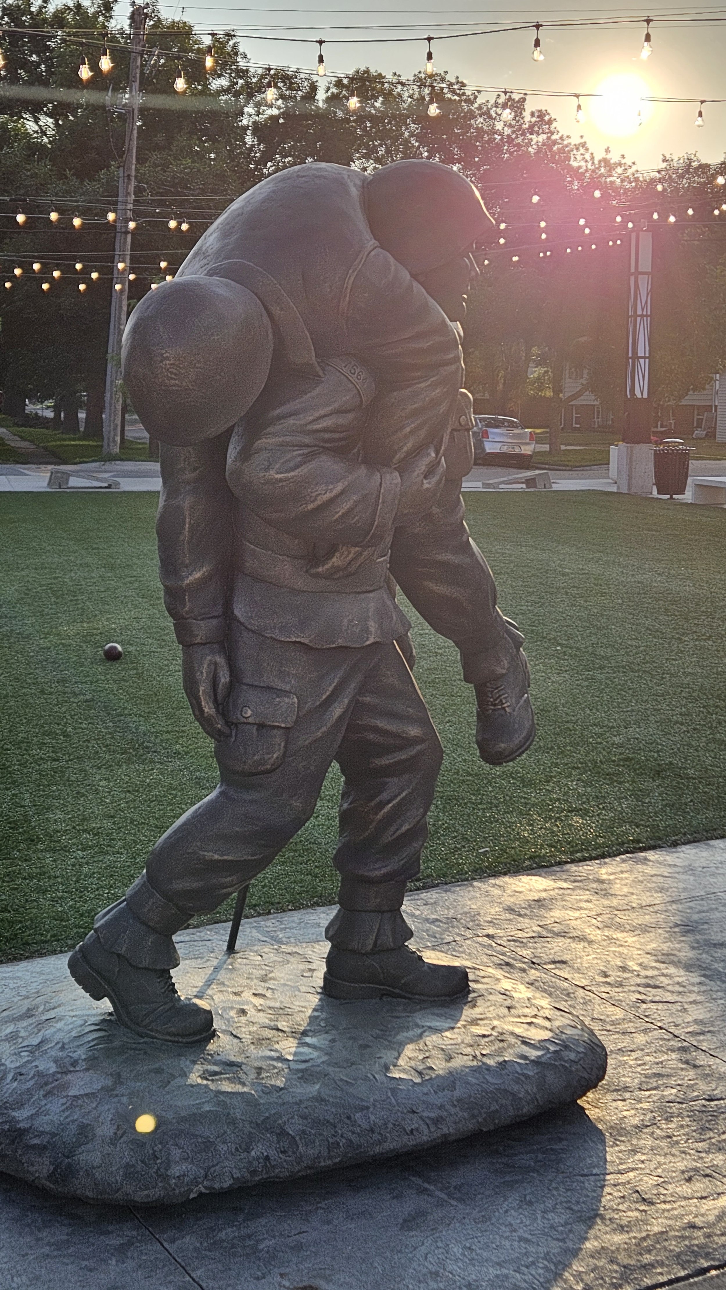Bronze statue of a soldier carrying a child, located outdoors with string lights overhead and a sunset in the background.