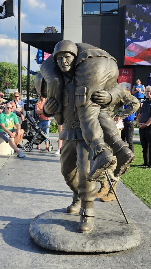 Bronze statue of a soldier carrying a wounded comrade, located outdoors with people and a modern building in the background.
