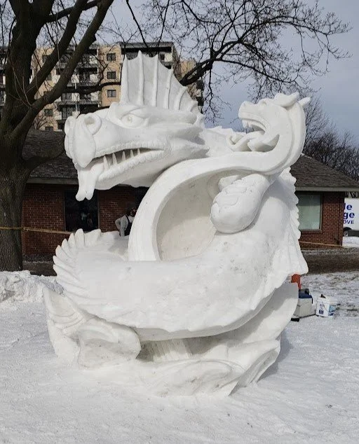 Sand sculpture of a dragon in a snowy outdoor setting, with buildings and trees in the background.