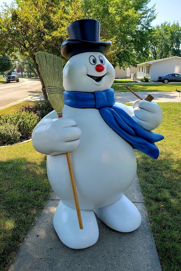 Large snowman statue with a black top hat, blue scarf, holding a broomstick and a shovel, standing on a sidewalk in a suburban neighborhood.