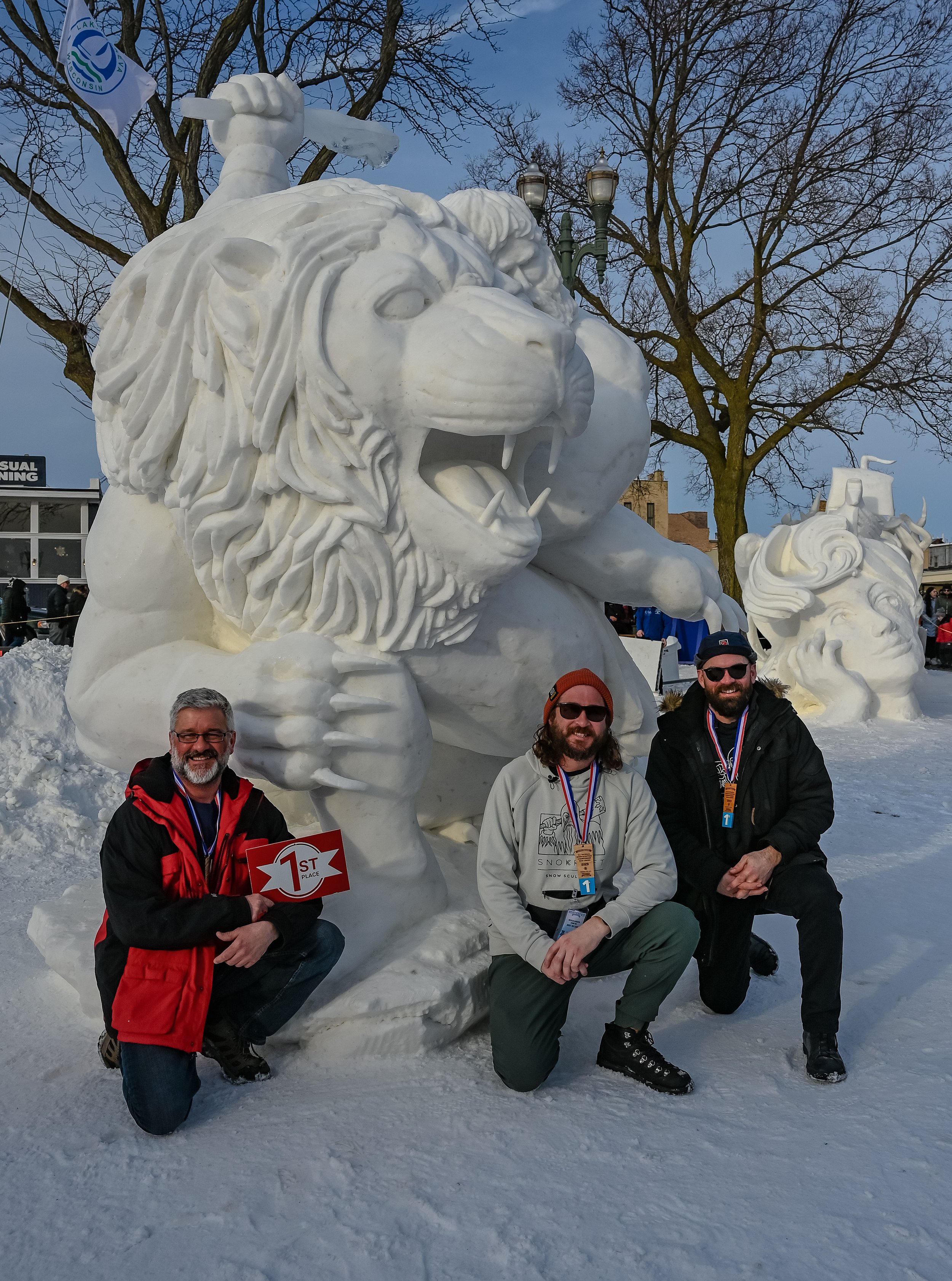Three men kneeling in front of a large snow sculpture of a lion with a mane, holding a first place sign. The sculpture is outdoors on snowy ground with leafless trees and street lamps in the background.