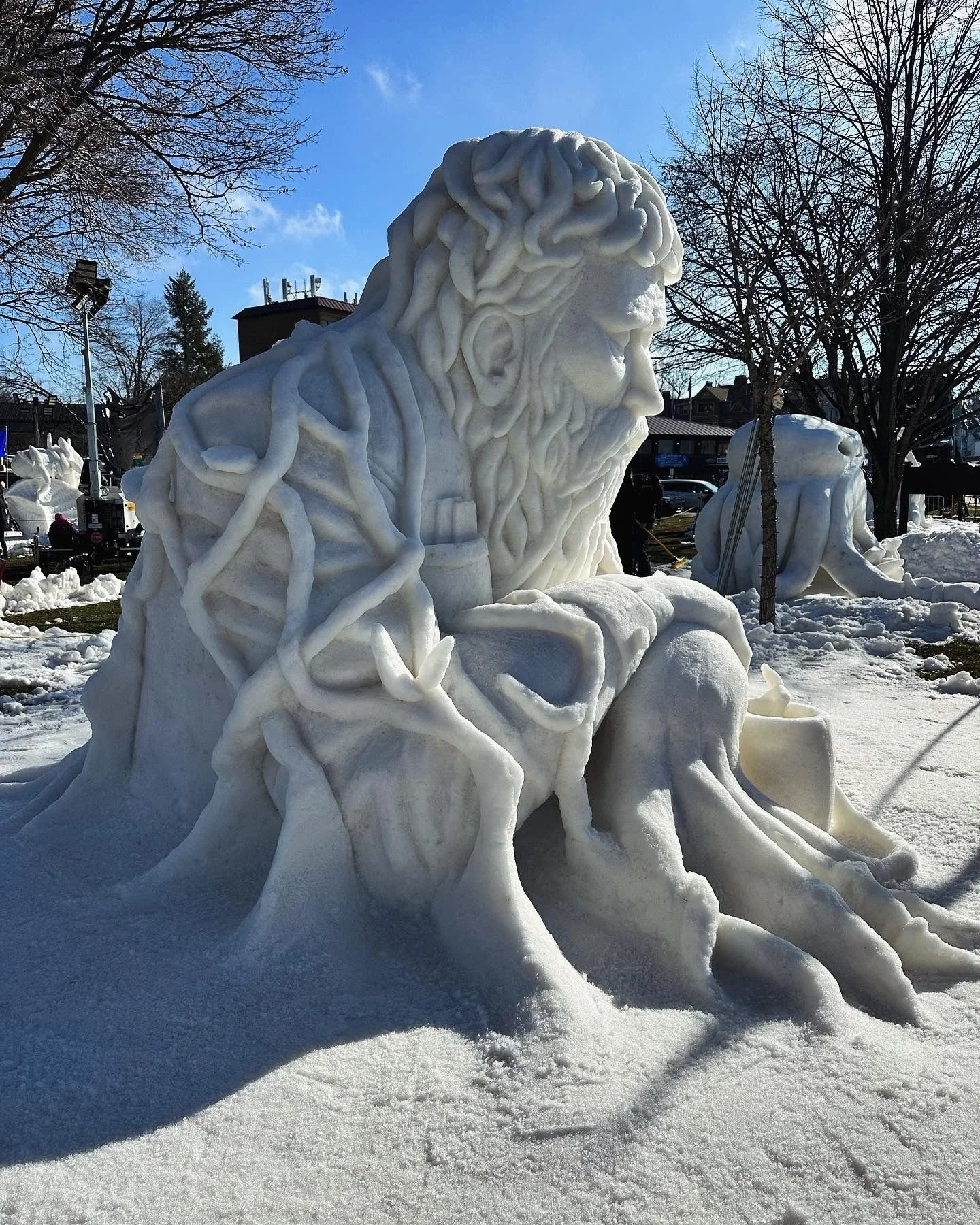 An intricate snow sculpture of a seated man with a beard and long hair, surrounded by snow formations and trees, with clear blue sky in the background.