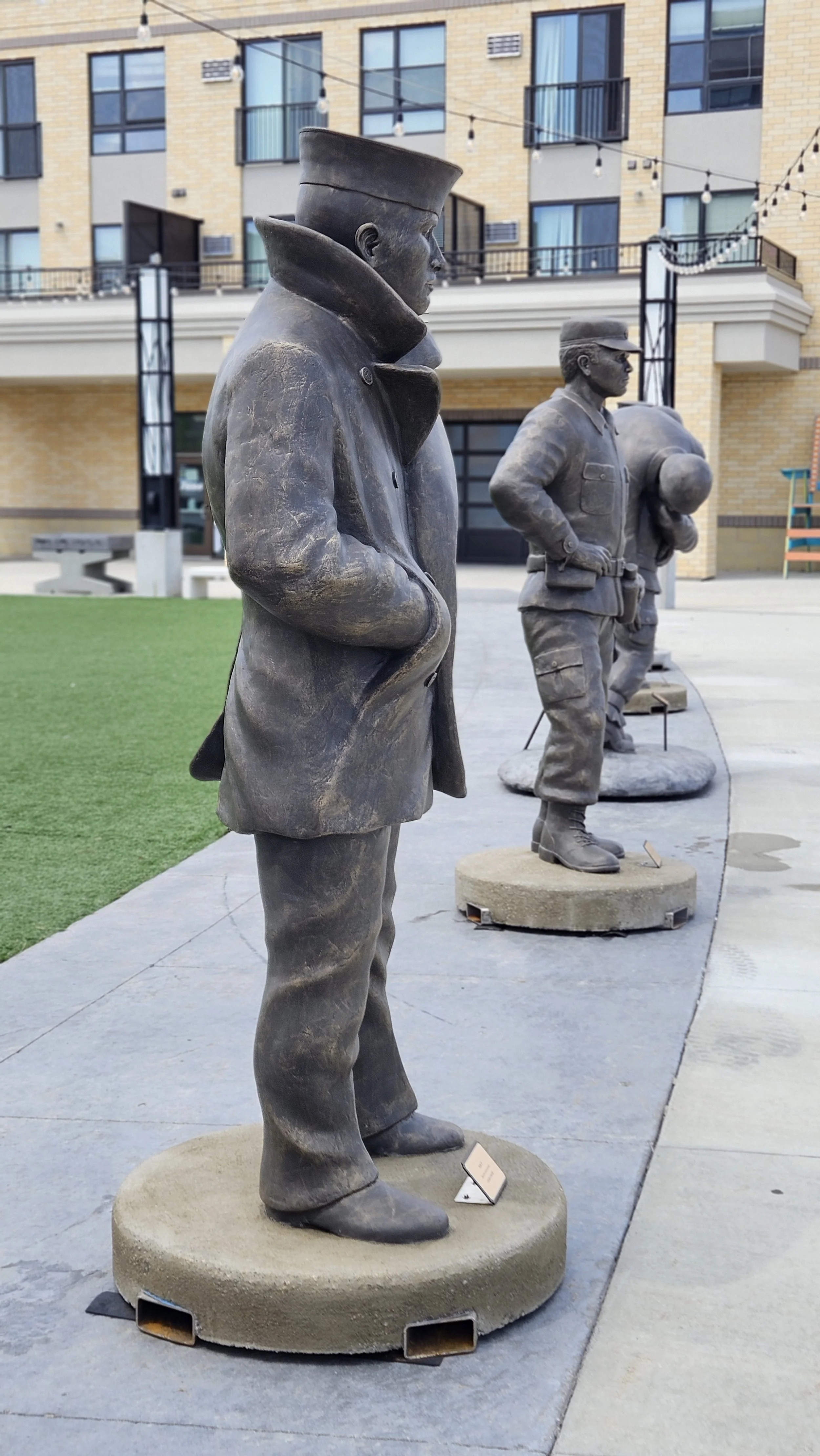 Bronze statues of soldiers in uniform, standing on round concrete bases, displayed outdoors in front of a modern residential building with balconies and windows.