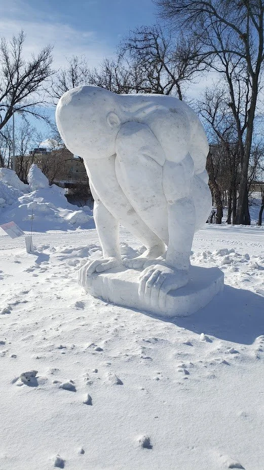 Snow sculpture of a person crouching with their head resting in their hands outdoors in a snowy landscape with leafless trees and a clear blue sky.