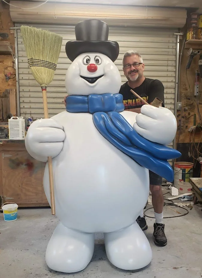 A man standing next to a large snowman sculpture in a workshop. The snowman has a black top hat, a red nose, a blue scarf, and is holding a broom. The snowman is smiling and giving a thumbs-up.
