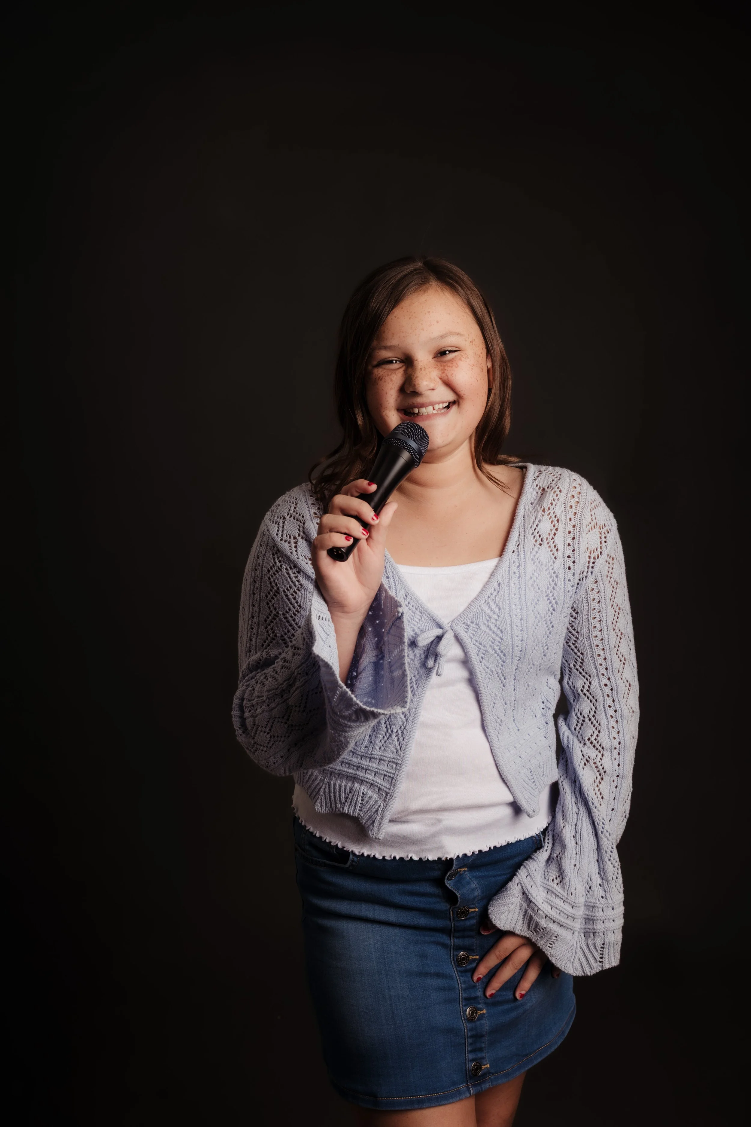 A young girl smiling and holding a microphone against a dark background.